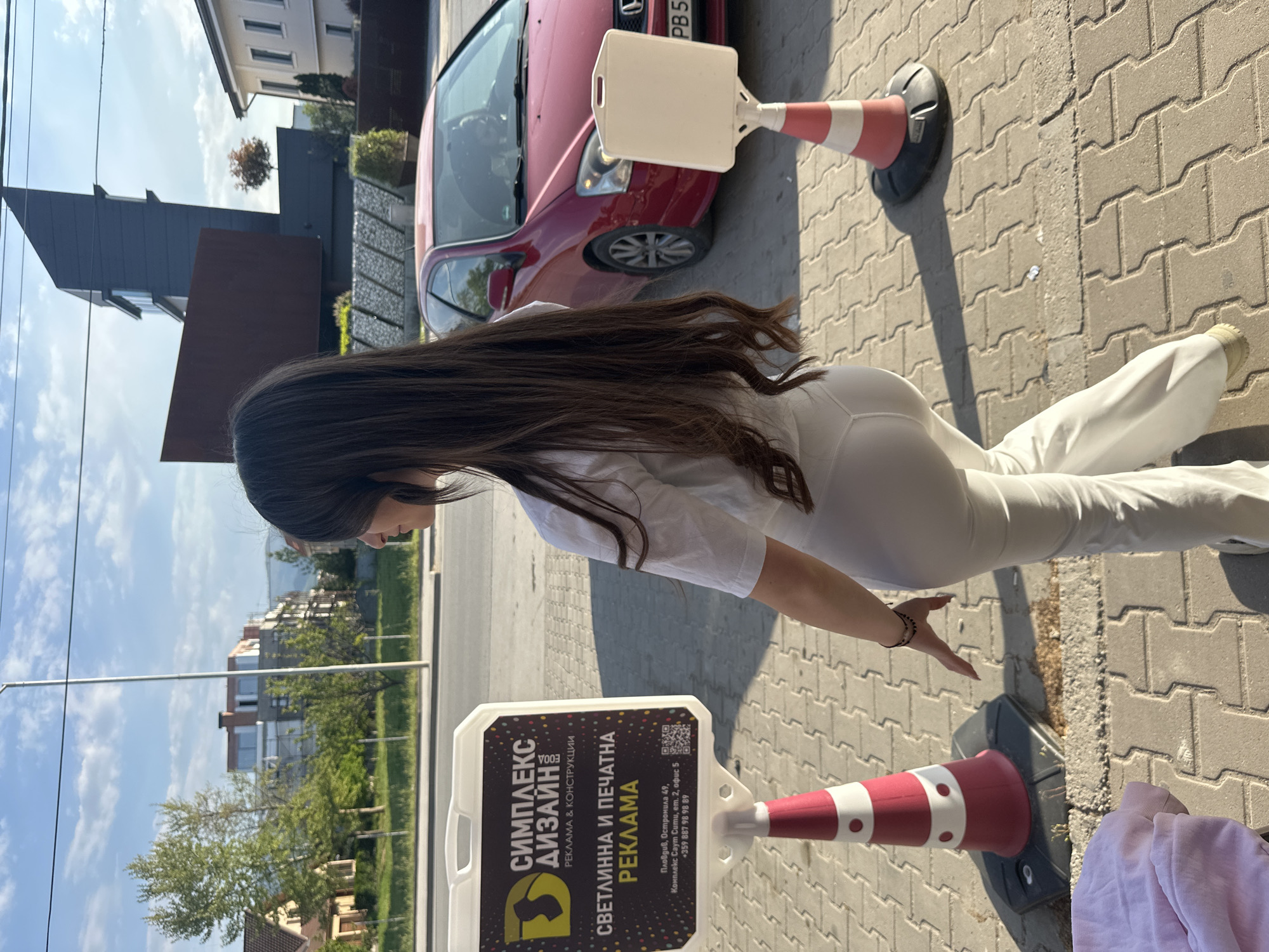 The image depicts an outdoor daytime scene on a paved sidewalk, likely in Plovdiv, Bulgaria. A person with long, dark hair, dressed in a white top and white pants, is standing beside a red and white advertising A-frame sign. The person is viewed from the back and side, with their head turned towards the left.
In the background, a red car is parked on the street, and various modern buildings, trees, and greenery are visible under a bright blue sky with some white clouds. The sun is high, casting distinct shadows on the pavement. Another red and white traffic cone, similar to the base of the A-frame sign, is visible further down the sidewalk.
The prominent A-frame sign displays text in Bulgarian, promoting an advertising and construction business. The text reads:
- "СИМПЛЕКС ДИЗАЙН" (SIMPLEX DESIGN)
- "РЕКЛАМА & КОНСТРУКЦИИ" (ADVERTISING & CONSTRUCTIONS)
- "СВЕТЛИННА И ПЕЧАТНА РЕКЛАМА" (LIGHT AND PRINT ADVERTISING)
- Contact information: "Пловдив, Остромила 49, Комплекс Саут Сити, ет. 2, офис 5" (Plovdiv, Ostromila 49, Complex South City, fl. 2, office 5)
- Phone number: "+359 887 98 98 89"
Two QR codes are also present on the sign. Part of a car license plate, "PB 5", is visible on the red car.