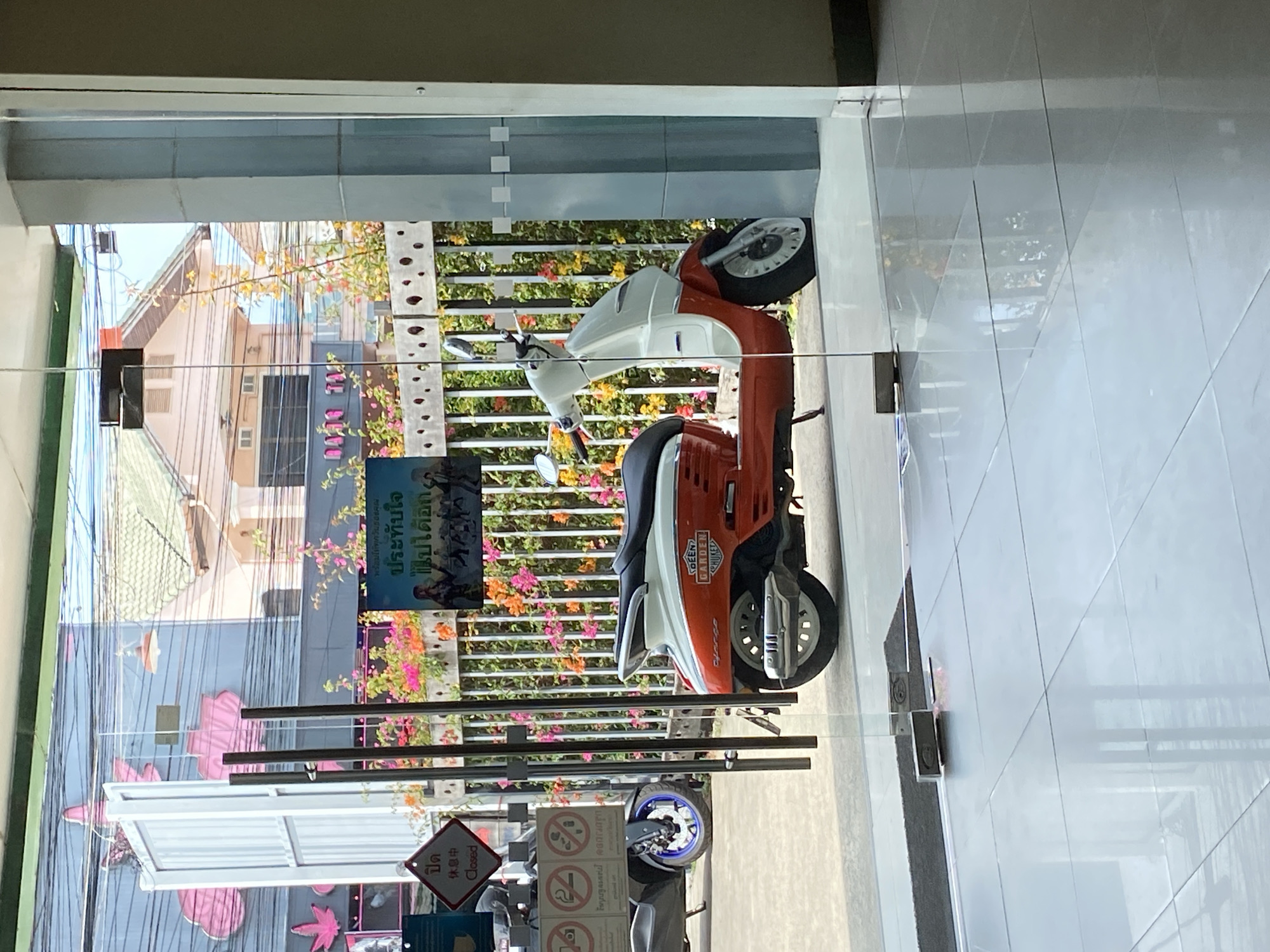 The image shows an orange and white scooter parked on the tiled floor of a building in Choeng Thale, Thailand. The scooter is reflected in a glass door or window. Outside, a street scene with buildings, a sign in Thai, flowers, and power lines is visible. The surroundings appear to be a commercial area. The overall lighting suggests a sunny day.