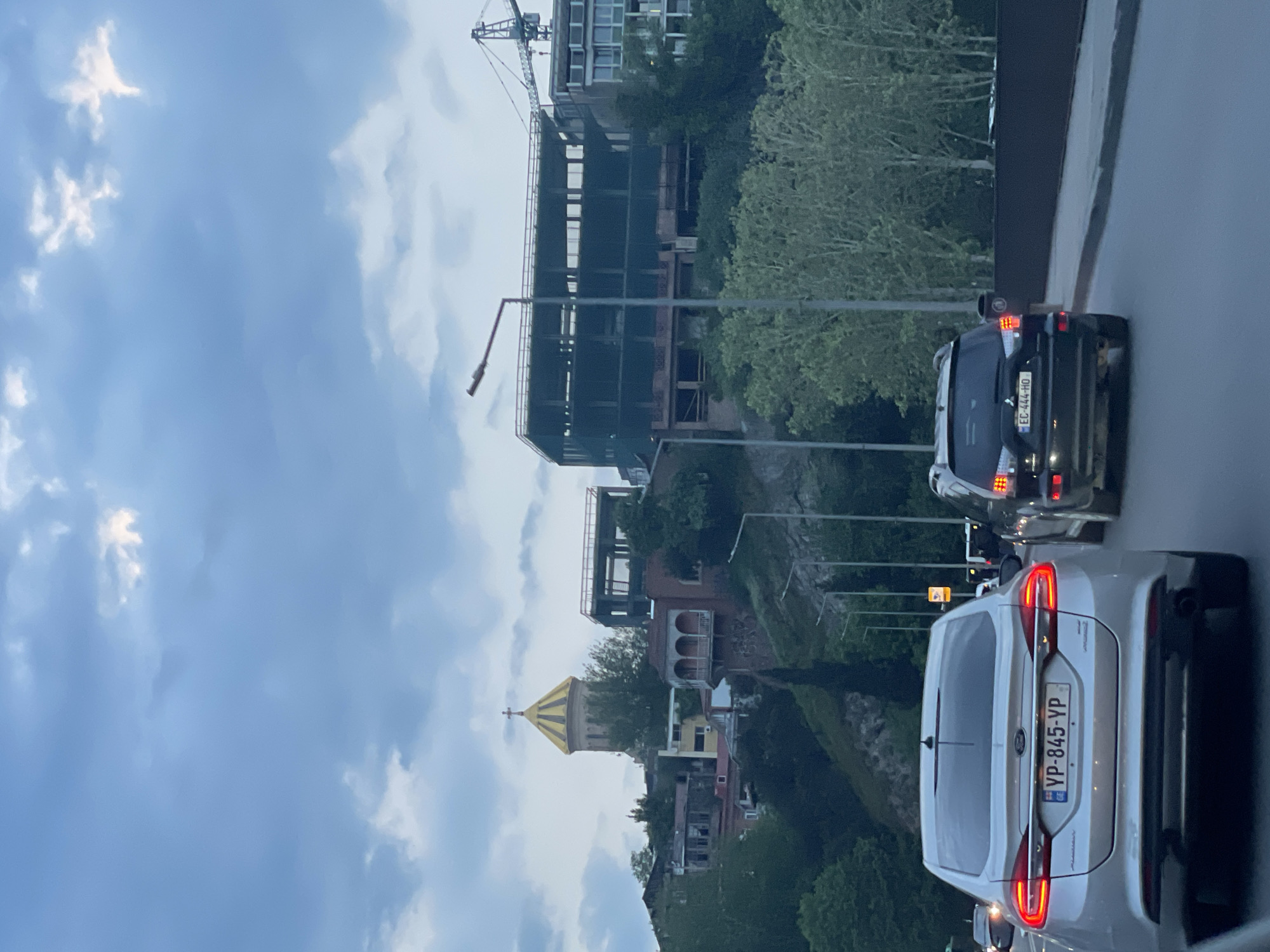 The image depicts a street scene in Tbilisi, Georgia, with a view overlooking a hillside with buildings, construction, and overcast sky.
Foreground: Two cars are visible on a road. A silver Ford station wagon with license plate "YP-845-YP" is closest to the viewer. Behind it is a dark SUV with license plate "EC-444-HO."
Midground: The scene overlooks a hillside with buildings and trees. A building under construction is visible on top of the hillside, with a crane extending above it. Further down the hill, a structure with a yellow conical roof and a cross on top is visible. Other buildings and trees are interspersed along the hillside.
Background: The sky is overcast with clouds.
Overall Impression: The image captures a common urban scene in Tbilisi, blending modern construction with older architectural elements against a cloudy sky.