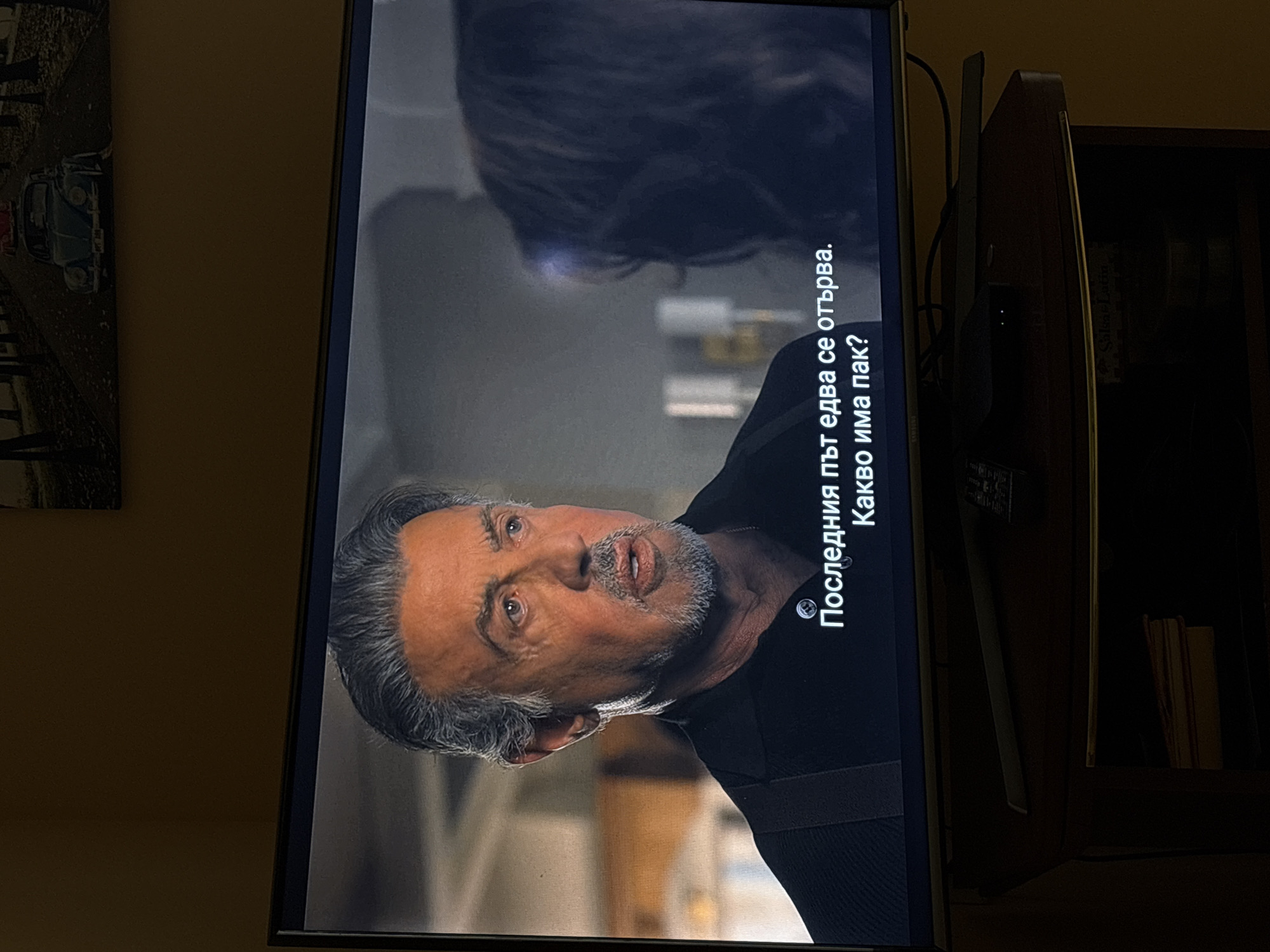 The image shows a television screen displaying a scene with subtitles in Bulgarian. The scene features a close-up of Sylvester Stallone, a Caucasian male with graying hair and a goatee, wearing a black collared shirt. He appears to be speaking. In the background, slightly out of focus, is another person, whose face is not visible, and an interior setting with visible light fixtures and furniture. The subtitles read "Последния път едва се отърва. Какво има пак?". This translates to "The last time he barely got away. What happened again?". The television is placed on a dark wooden TV stand. The background wall is a solid, warm, light brown color. There is a framed picture on the wall above the television, depicting a street scene with a vintage car. There are no visual elements in the image that indicate the location is the city of Provadia, Bulgaria.