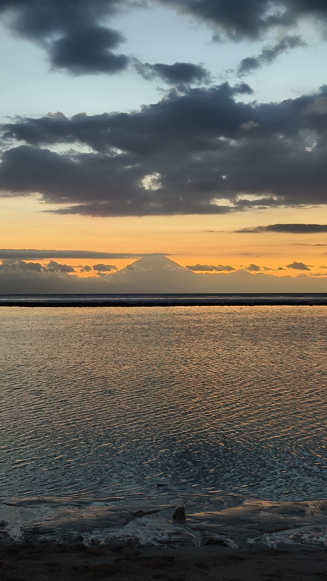 The image shows a body of water with small ripples reflecting the warm tones of the sky at dusk or dawn. A faint mountain silhouette is visible on the horizon, partially obscured by a layer of clouds. Above, heavier, darker clouds fill the upper portion of the frame. The foreground depicts a beach with some ice, possibly a tidal flat, indicating the water is shallow near the shore. The scene is generally calm and subdued in lighting. The city is not apparent in the image. Indonesia is specified as the location.
