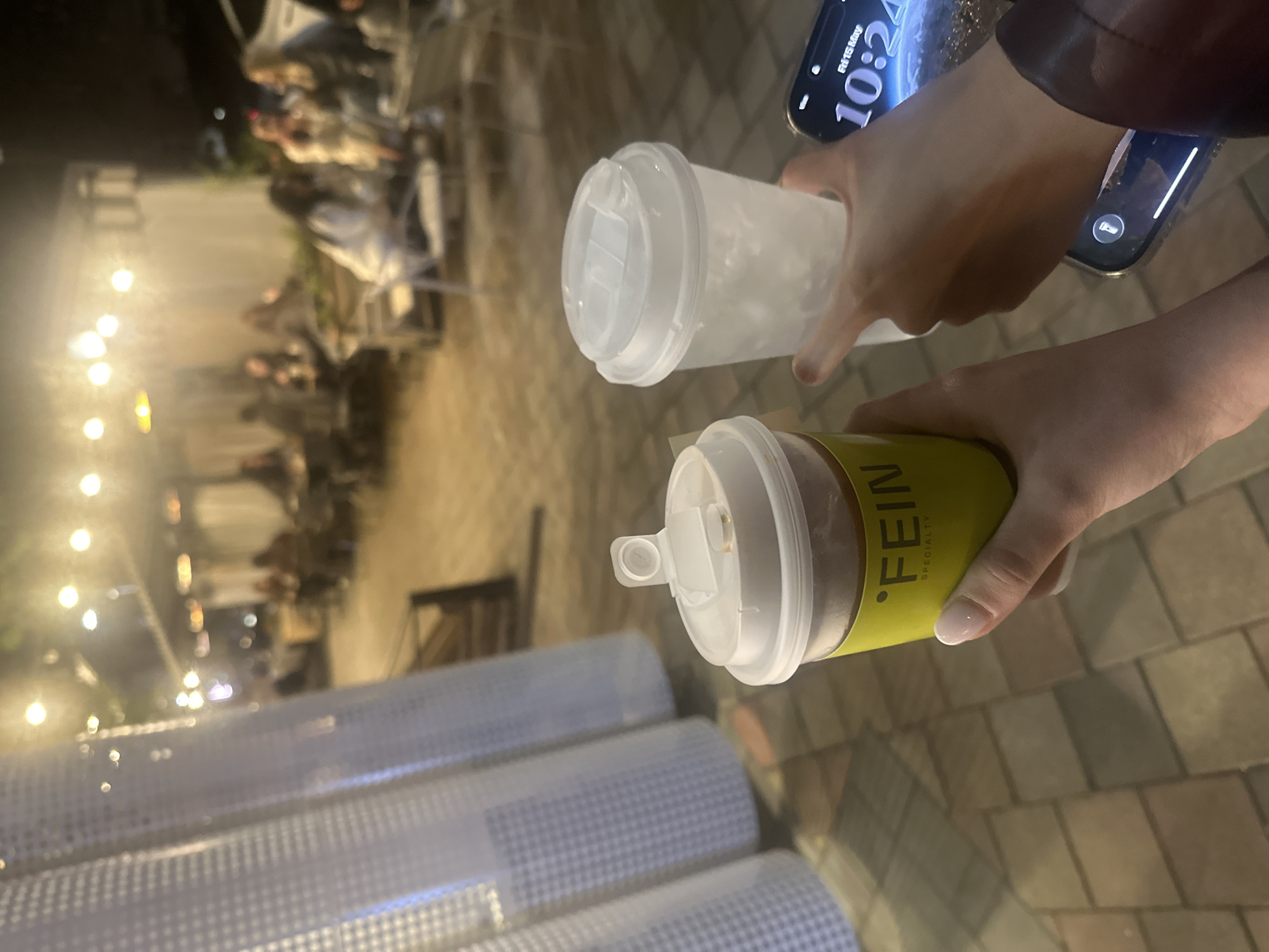 The image shows two people holding drinks outside at night. One person holds a clear plastic cup with a white lid, and the other holds a yellow-labeled cup with the word "FEIN" visible, also with a white lid. The person with the clear cup also holds a phone displaying a time of 10:24 AM and the date "Fri 15 May." The background features a blurred view of an outdoor seating area with people and illuminated lights, suggesting a restaurant or cafe setting. Rolls of plastic netting are stacked in the lower part of the frame. The location appears to be in Yerevan, Armenia, based on the “FEIN” brand on one of the cups. The primary action involves holding and potentially consuming drinks at an outdoor venue in the evening.