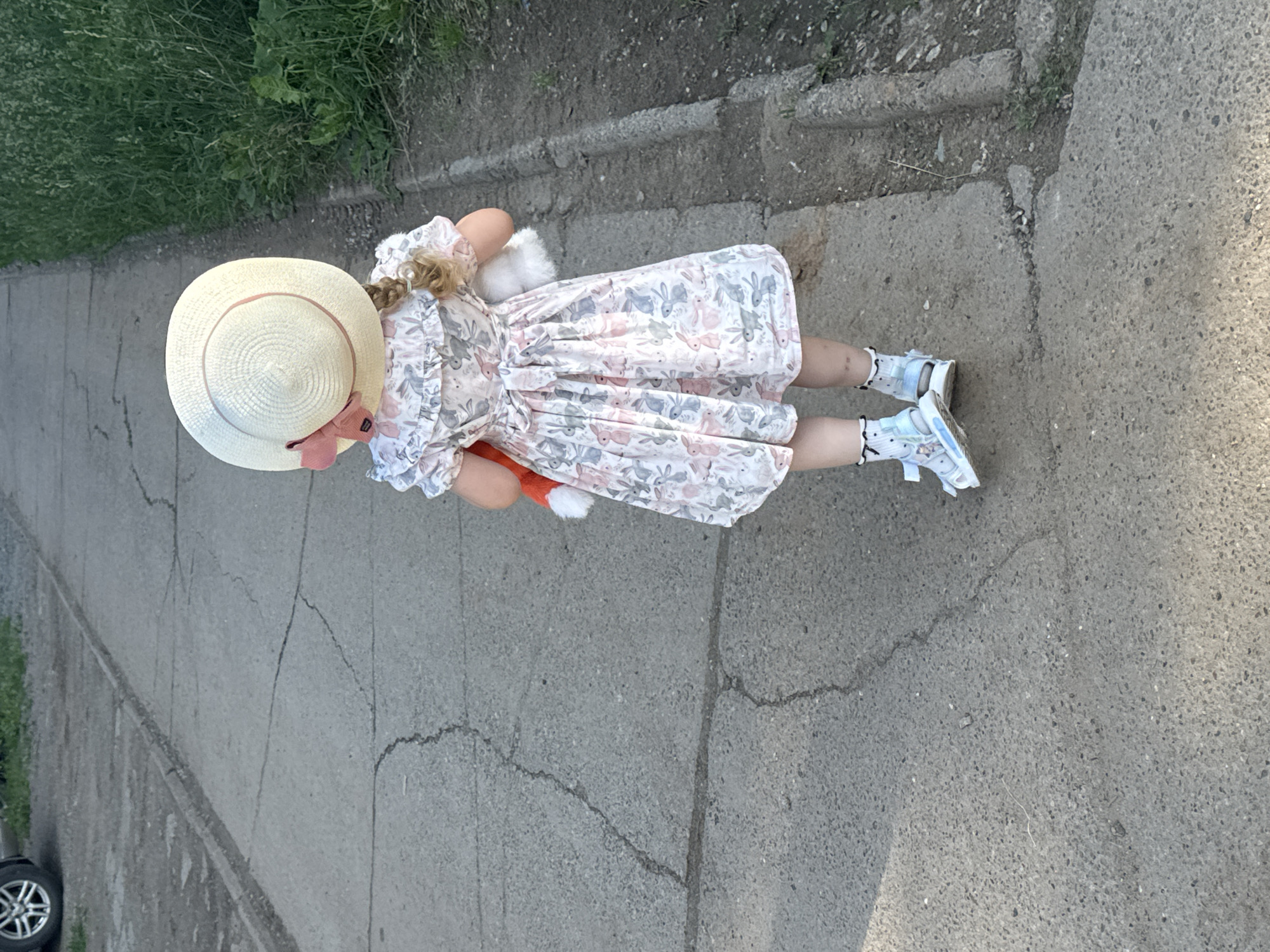 A young girl, viewed from behind, walks on a cracked asphalt path in Chaikovsky, Russia. She wears a white straw hat with a pink ribbon, a light-colored dress with a rabbit pattern, frilly socks, and blue and white sandals. She holds a stuffed orange fox in her left arm and a white stuffed animal in her right arm. The path is bordered by a grassy area on the right and a dirt and gravel area on the left, where a car wheel is partially visible. The scene appears to be outdoors in daylight.