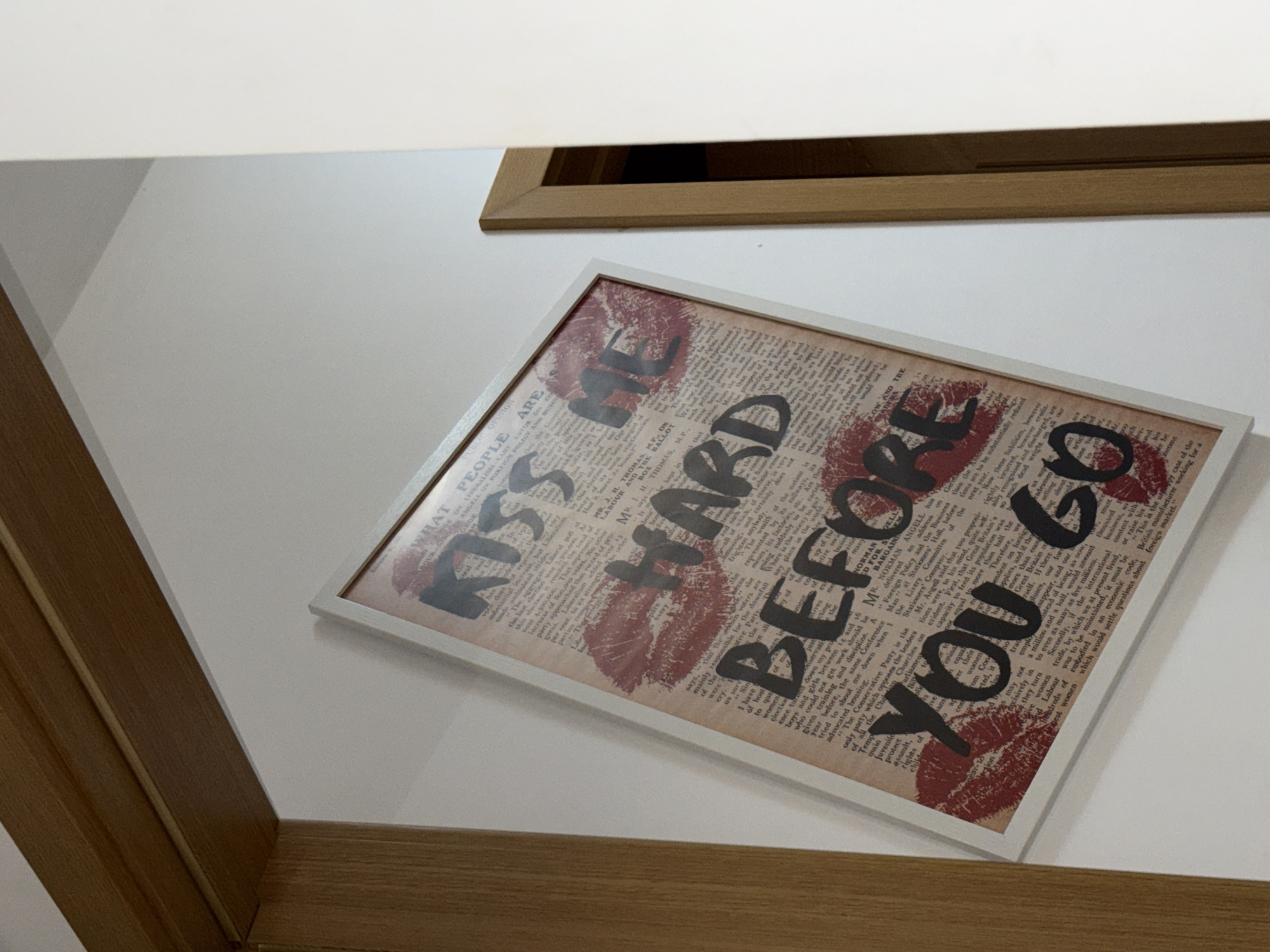 A framed print with the words "Kiss Me Hard Before You Go" is hanging on a white wall. The print is styled to look like a newspaper with lipstick kiss marks. A wooden frame borders the wall and print. Another wooden frame is partially visible above the print. The location is unknown; there is no information to indicate that this image was taken in Plovdiv, Bulgaria.