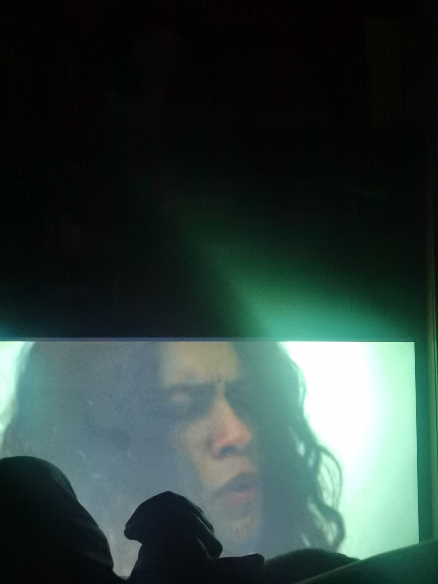A close-up shot shows a person with long, dark, wavy hair and visible facial features (nose and mouth) on a television screen. The screen is emitting a bright, greenish light. Below the screen, in the foreground, are dark, indistinct shapes that appear to be part of a person sitting in front of the television. The background above the screen is dark. The image contains no visible information to determine a location in Chaikovsky, Russia.