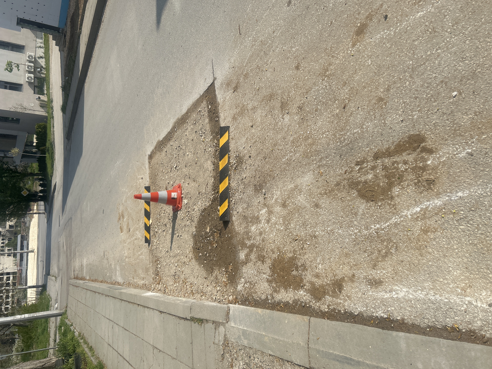 The image shows a section of road in Pleven, Bulgaria, undergoing repair. A square area has been excavated from the road surface, revealing a gravelly base. A traffic cone and two striped bollards are positioned around the excavation to alert pedestrians and vehicles. The road surface is asphalt. A concrete curb is visible along the side of the road. In the background, buildings are visible. There are no people in the image.