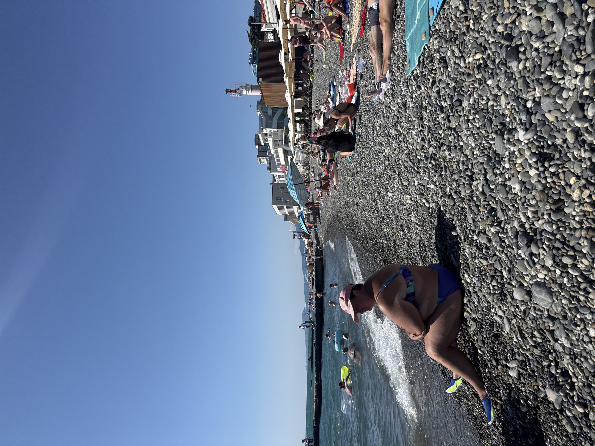 A wide shot captures a sunny day at a crowded pebble beach in Sochi, Russia. In the foreground, a woman in a pink hat and a blue and purple bikini sits on the pebbles, facing the ocean. The water is a deep blue with gentle waves lapping at the shore. Several people are visible in the water, some floating on inflatable rings.
The beach is packed with people sunbathing on towels and loungers. Umbrellas are set up, providing shade. In the mid-ground, buildings and a lighthouse are visible in the distance, suggesting an urban setting. The sky is clear and blue. The overall scene depicts a lively beach resort on a warm day.