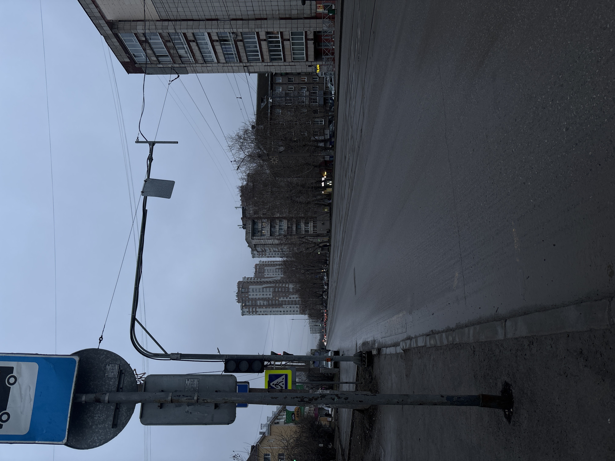 The image shows a street scene in Perm, Russia. The view is looking down a wet, paved road lined with buildings of varying heights and architectural styles. The sky is overcast, suggesting a dull or potentially rainy day. A traffic light and several street signs are visible in the foreground, with the prominent blue and white sign partially obscuring the view. Bare trees line the road, hinting at a cold season. Multiple utility poles and wires run overhead, adding to the urban landscape. In the distance, there is a line of vehicles along the road, which indicates traffic.