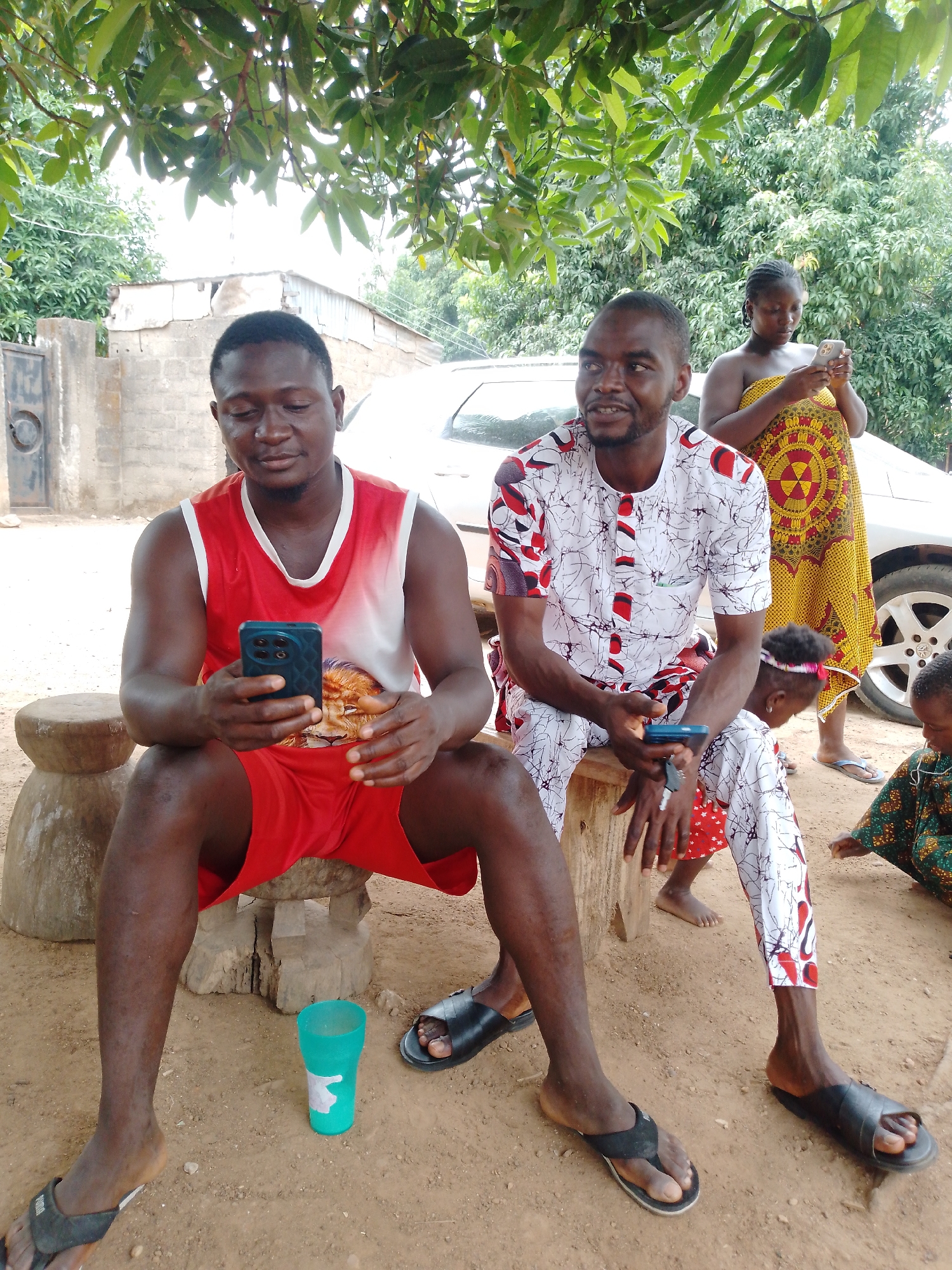 The image shows a group of people outdoors. Two men are seated on wooden stools, both looking at smartphones. The man on the left wears a red and white tank top and red shorts, while the man on the right is wearing a white patterned shirt and pants. A woman stands behind them in a yellow and red patterned dress, also using a smartphone. Two children are present near the seated man on the right, one with a headband and the other in a green patterned outfit. A white car is visible in the background, and a green plastic cup sits on the ground. The environment appears to be a residential area with trees and a building in the background. The location is the city of Unknown, Unknown.