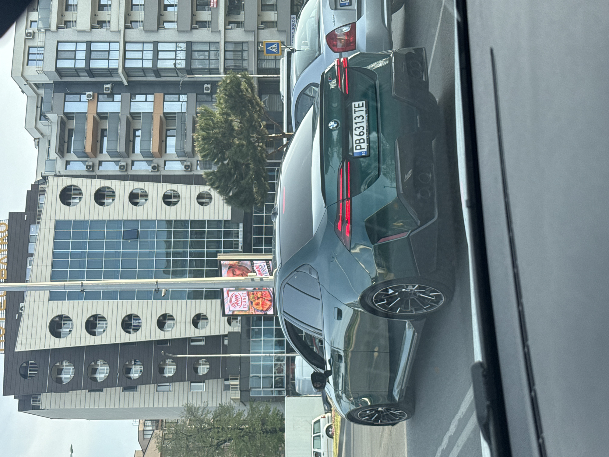The image depicts a dark green BMW car with a Bulgarian license plate "PB6313TE" in Plovdiv, Bulgaria, as evidenced by the license plate code "PB." The car is situated on a street with buildings in the background, notably a modern building with circular windows. Other vehicles are present nearby. The photo appears to have been taken from inside another vehicle. There is a billboard in the background with food advertisement.