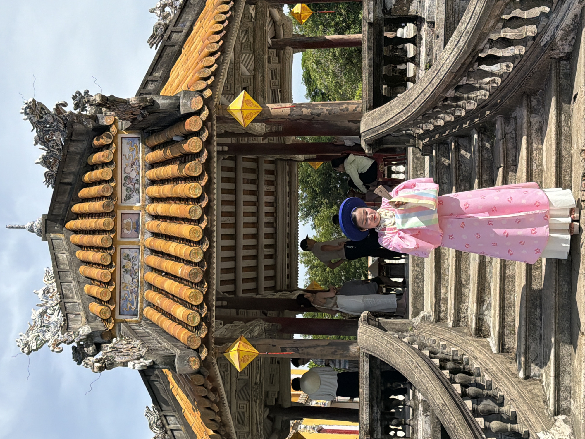 A young woman in traditional pink clothing stands on a stone staircase in front of a pavilion with an orange tiled roof in Phú Xuân Ward, Vietnam. The pavilion's roof is decorated with ornate dragon figures and colorful ceramic tiles. Yellow lanterns hang from the pavilion's beams. Several other people are visible in the background, some standing and others seated. The staircase has weathered stone railings and appears to be part of an ancient structure. The woman is holding a colorful striped bag and looking down at a piece of paper in her hands. The sky is partly cloudy.