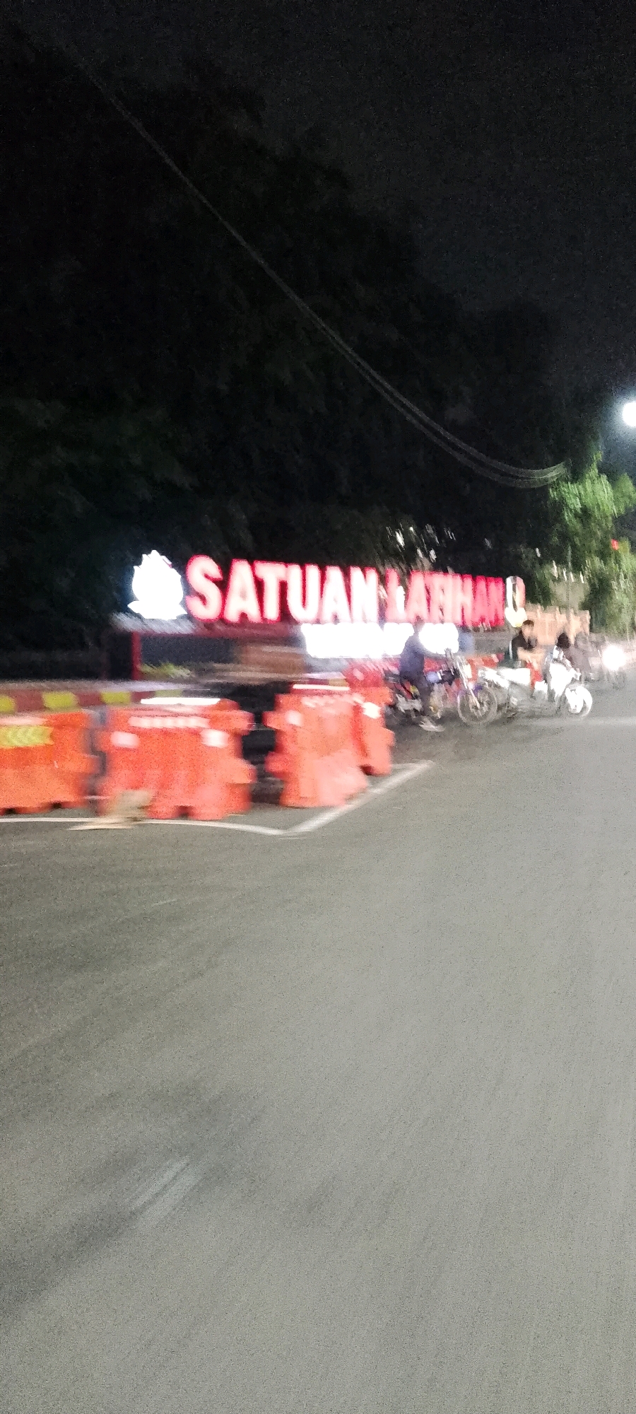 A blurred, nighttime photograph shows a street scene in Gunung Putri, Indonesia, with a prominent illuminated sign reading "SATUAN LATIHAN" in red lettering. The sign is partially obscured by orange traffic barriers and a row of trees in the background. Several motorcycles and individuals are visible in the vicinity of the sign, suggesting activity or traffic. The road surface appears to be asphalt. The overall impression is one of a busy intersection or a facility entrance at night.