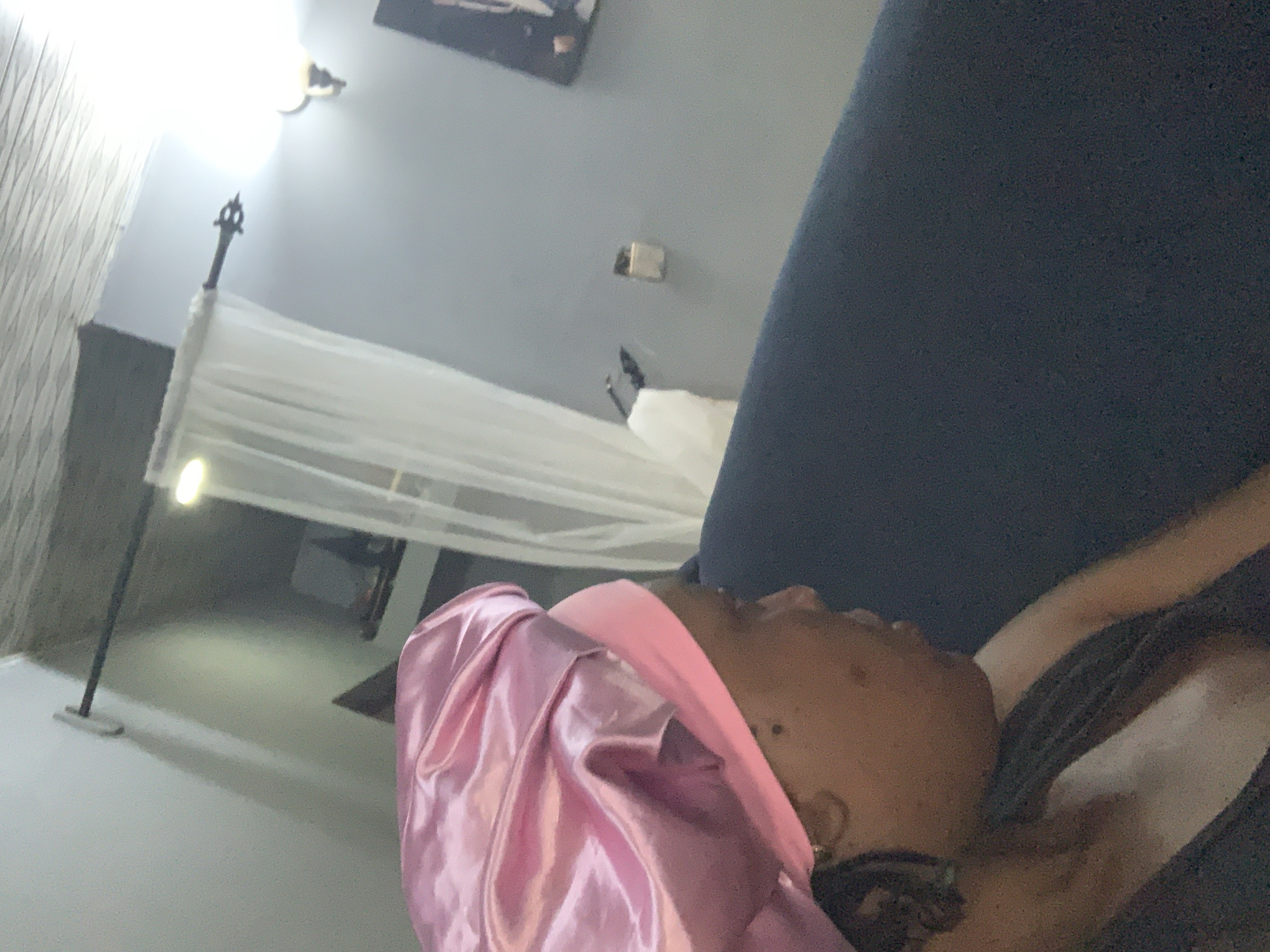 The image shows a person wearing a pink satin hair bonnet, reclining against a dark blue sofa. A sheer white canopy hangs from a tall, slender floor lamp behind the person. The room has light-colored walls. A framed picture is mounted on the wall. The location, Sabon Tasha, Nigeria, cannot be determined from the image alone.