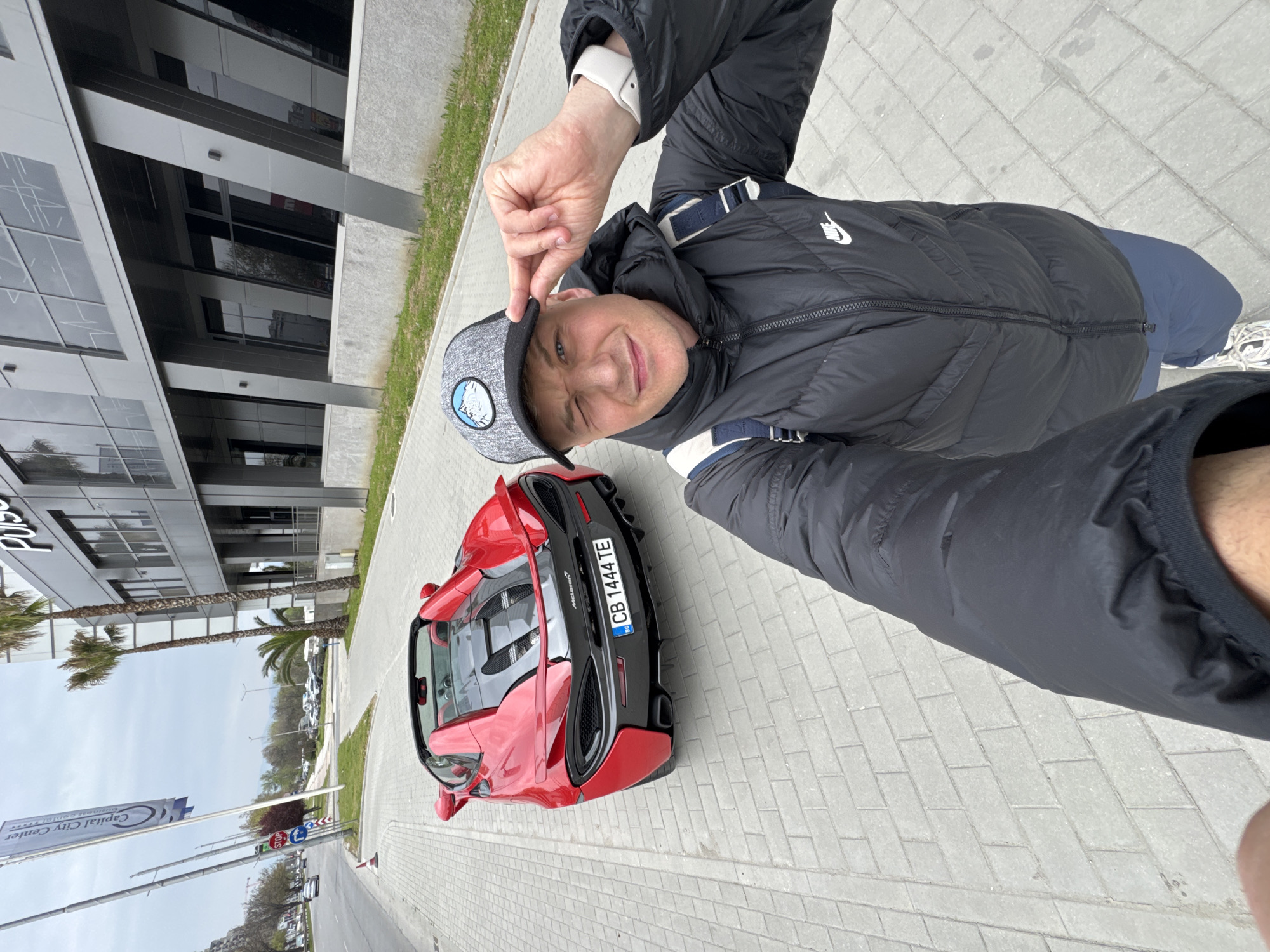 A man is taking a selfie next to a red McLaren sports car. He is wearing a black puffer jacket, a gray hat with a logo, and a white wristwatch. The car has a Bulgarian license plate that reads "CB 1444 TE". The background shows a modern building with glass windows and palm trees. A sign that reads "Capital City Center" can be seen. The location appears to be in Plovdiv, Bulgaria. The scene is set on a paved walkway next to a street.