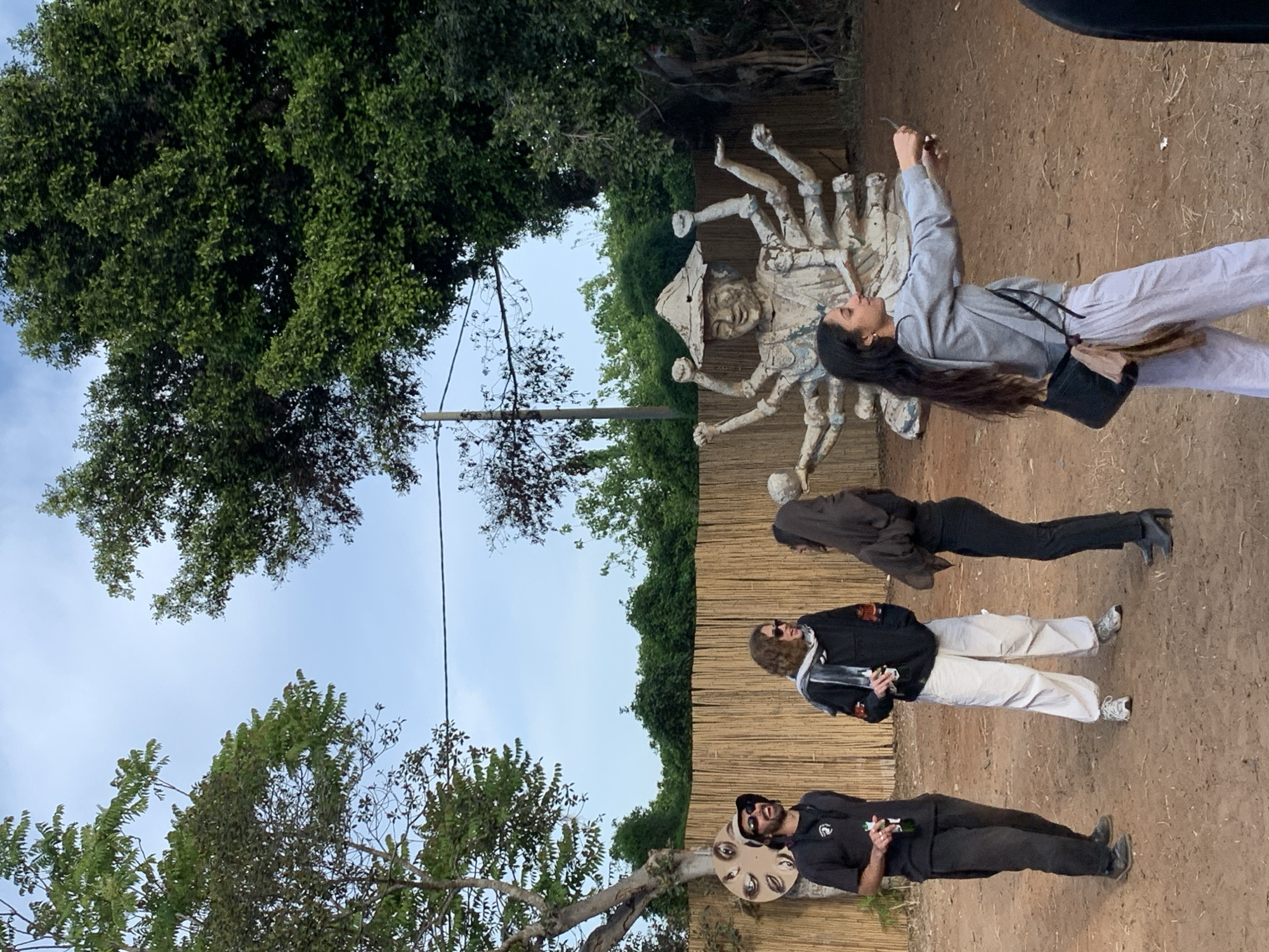 The image shows a group of four people in what appears to be an outdoor art installation or sculpture garden, potentially in Gan Soreq, Israel. One woman is posing, holding up a cell phone, next to a large, light-colored sculpture of a figure with multiple arms and a conical hat. She wears a grey sweatshirt, white pants, and a black shoulder bag. A second woman, wearing a brown jacket and black pants, stands behind her. A man with sunglasses and white pants is positioned behind the second woman. The final man, at the front of the group, wears a black shirt and pants and holds what looks like a bottle. Near him is a disc shaped object with eyes on it. The background consists of a tall bamboo fence with lush green trees above and behind it and blue sky with some white clouds visible overhead. The ground is dirt.