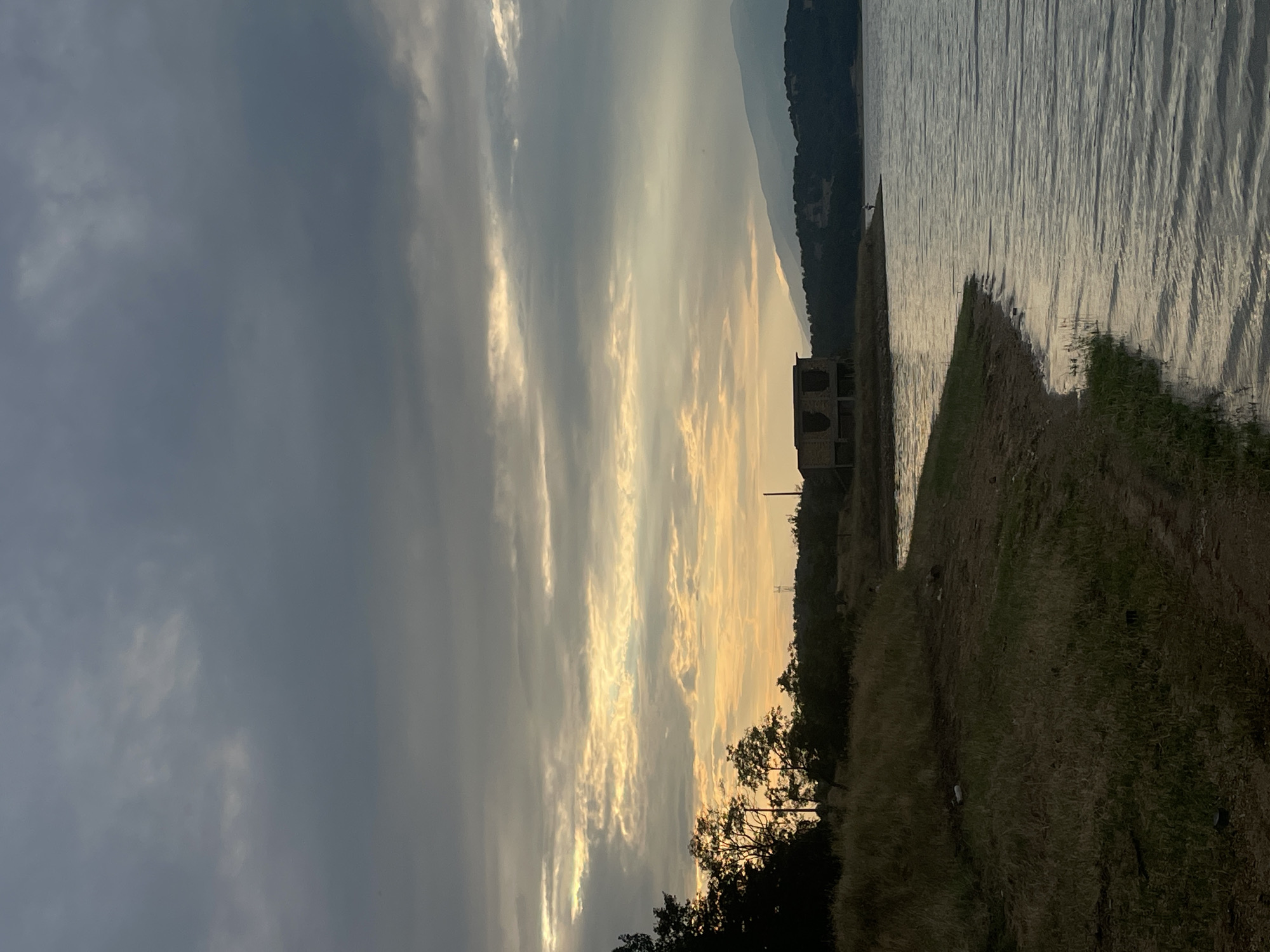 This is a vertical, eye-level, full shot of a serene lakeside landscape at dusk. The sky is dramatic, with dark, heavy clouds dominating the left side, interspersed with bright, golden streaks of sunlight breaking through on the right, creating a stark contrast. In the distance, the silhouette of a forested mountain range forms the horizon.
The foreground features a grassy embankment sloping down towards a calm body of water. The water reflects the muted colors of the sky, with subtle ripples suggesting a gentle breeze. On the embankment, several trees with dark foliage are visible, some appearing as silhouettes against the lighter sky. A portion of an old, stone building with arched openings stands on the right side of the frame, adding a touch of historical intrigue to the scene. The ground is covered in dry, yellowed grass, hinting at the end of summer or beginning of autumn.
The overall mood is peaceful and contemplative, amplified by the soft, fading light of sunset. The presence of the building suggests a past human presence, now reclaimed by nature. The lack of any visible people or explicit activity points towards a quiet, natural moment. The time of day is clearly evening, with the dramatic lighting and cloud formations characteristic of sunset. No text is visible in the image. The location is identified as Domlyan, Bulgaria, which aligns with the natural landscape depicted.