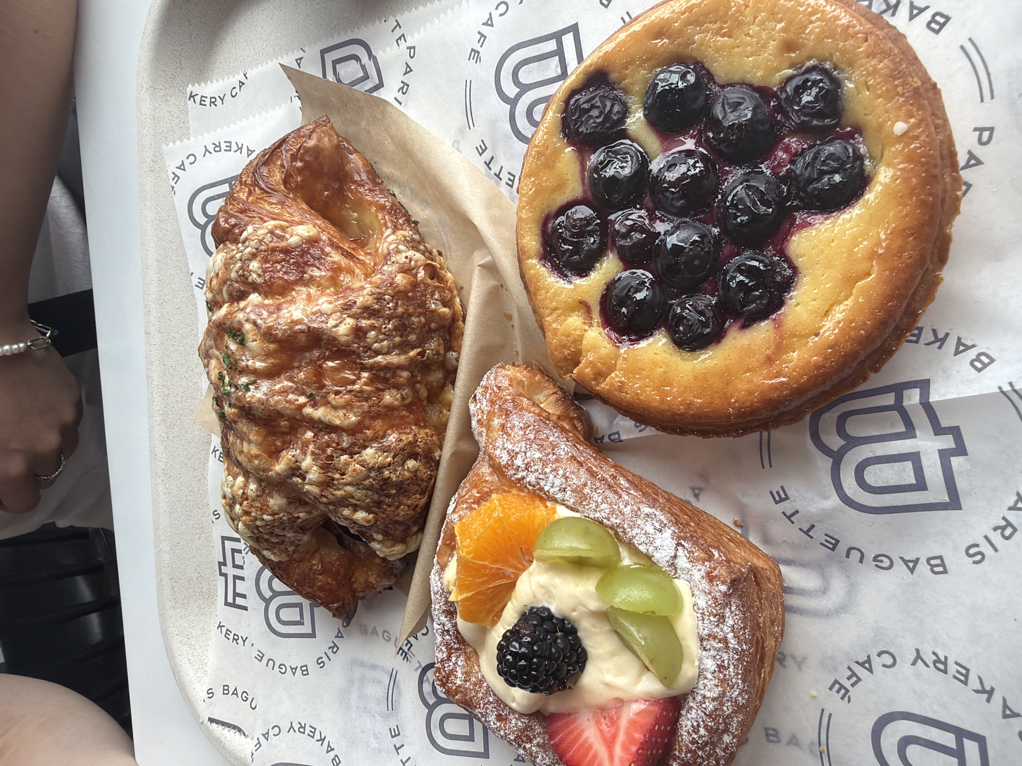 A close-up overhead shot shows three pastries on a white paper liner on a gray tray. The pastries are a savory croissant with melted cheese and herbs, a fruit danish with cream cheese, strawberries, blackberries, grapes, and orange slices, and a blueberry tart with a custard base. The paper liner has a repeating pattern of the word "PARIS BAGUETTE CAFE" and a stylized "B". The image appears to be taken indoors, with natural light illuminating the scene. The upper right corner of the image shows a partial view of two individuals, one with a silver ring on their left ring finger. The location is identified as Beaverton, United States of America.