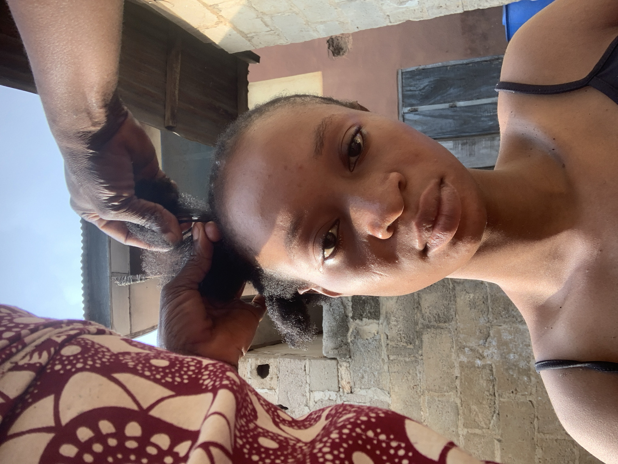 The image captures a close-up of a young Black person, facing the camera with a neutral expression. Their head is tilted, and their gaze is directed slightly off-camera. They appear to be outdoors, with the background showing a brick wall and what looks like a corrugated metal roof. The lighting suggests it's daytime, possibly late morning or early afternoon, with sunlight casting shadows.
The person's hair is being styled, with hands visible in the foreground working on their hair. The hands appear to be braiding or twisting the hair, which is dark and textured. The person is wearing a dark-colored top with a thin strap visible across their shoulder. A portion of another person's torso, wearing a patterned garment with maroon and cream colors, is also in the foreground, indicating they are likely the one styling the hair. The overall scene has a candid and intimate feel, suggesting a personal moment of hair care. The presence of the brick and metal structures hints at a rustic or residential setting, consistent with the provided location context of Shagamu, Nigeria. No visible text is present in the image.
