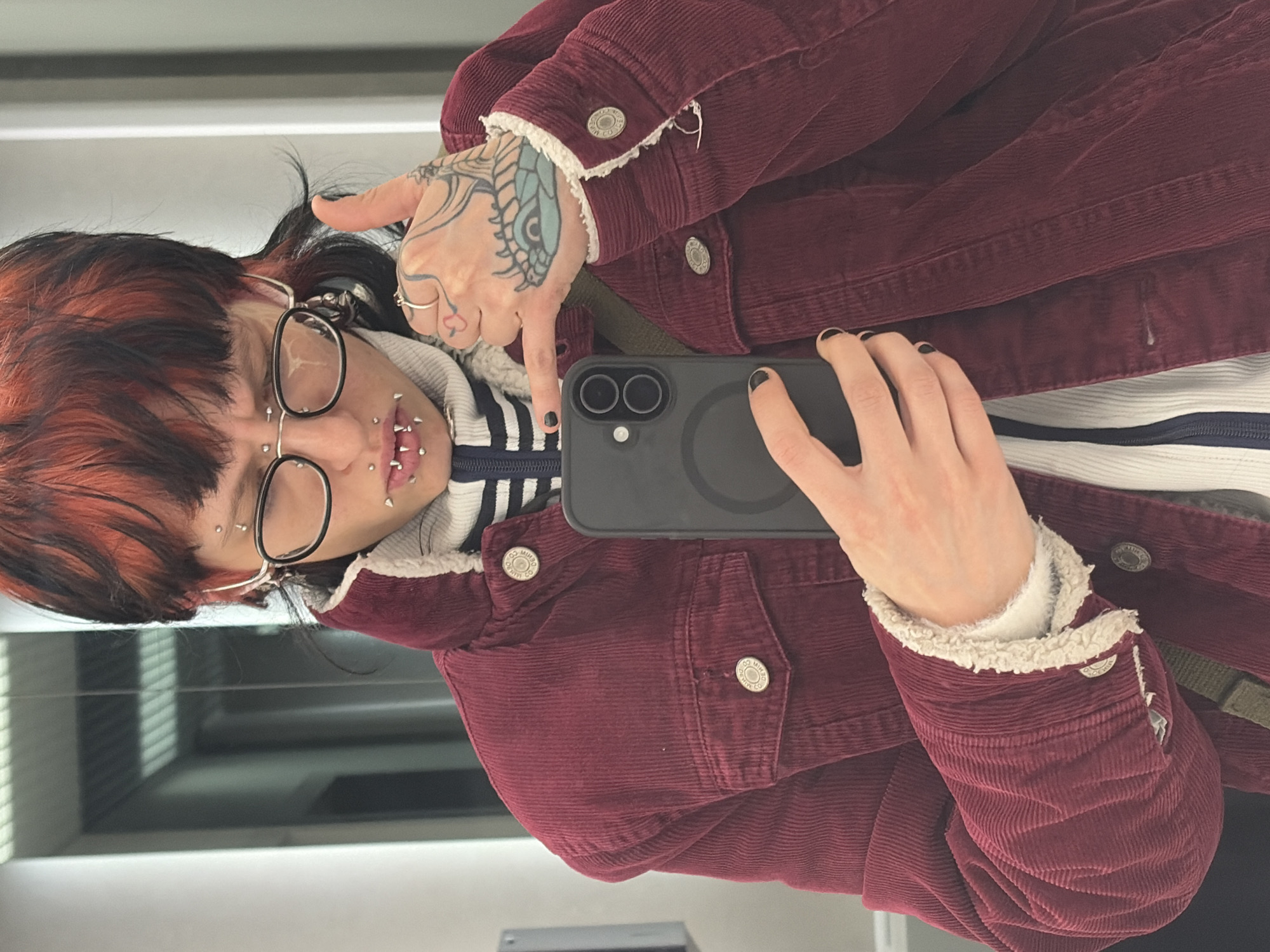 A person is shown from the chest up, holding a phone in their right hand in front of them. The person has visible facial piercings on their lip and eyebrow. They are wearing glasses and have red-streaked hair. Their left hand is visible, with a tattoo on the back of their hand. They are wearing a burgundy corduroy jacket with a white, sherpa-like lining, and a striped shirt underneath. The background is blurry, showing a window with blinds. There is no indication of this image being taken in Busmantsi, Bulgaria.