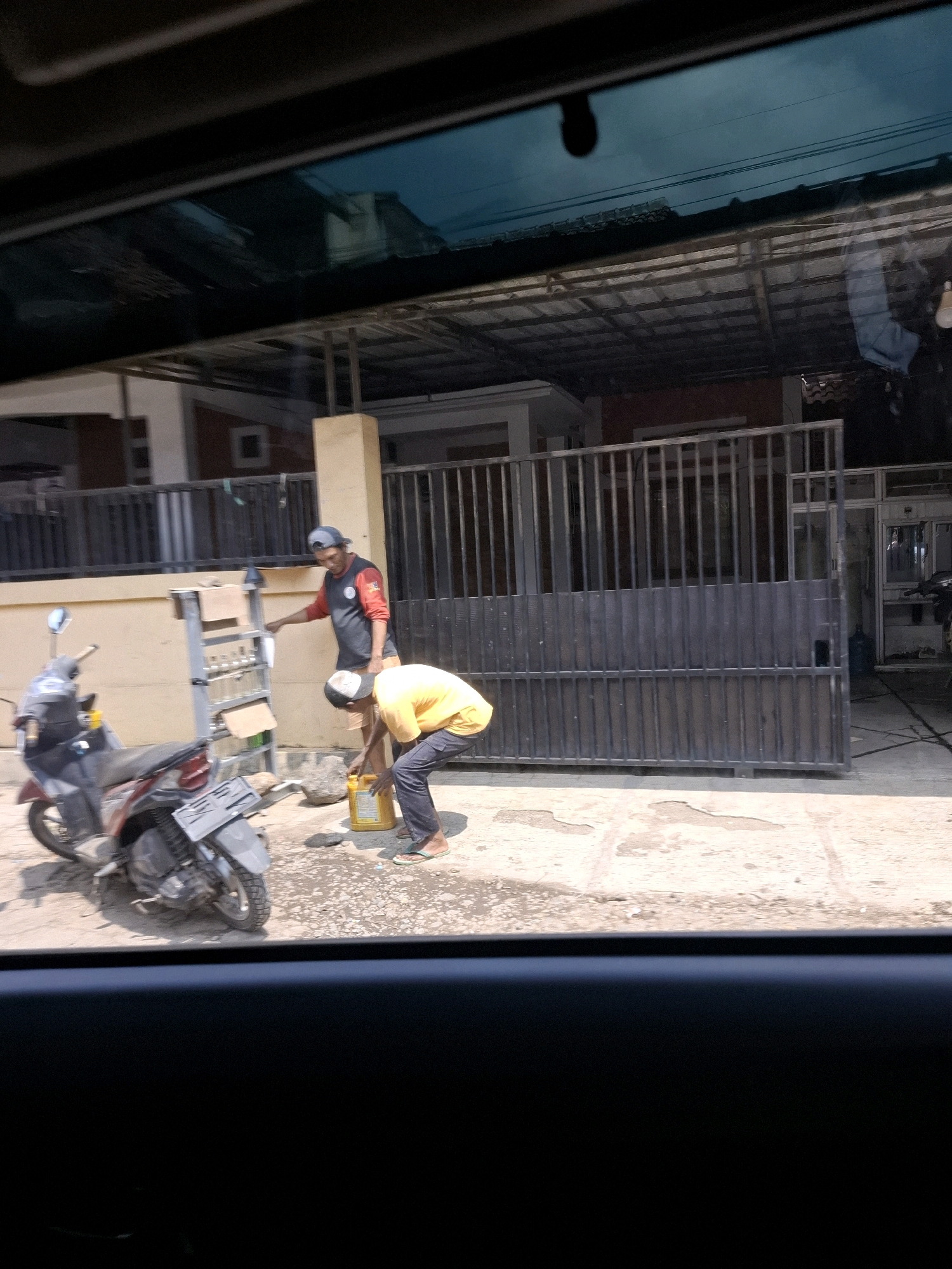 In Ciheulang Tonggoh, Indonesia, two individuals are visible on a paved area outside a building. One person, wearing a dark shirt with red sleeves and a baseball cap, stands near a silver ladder and gestures towards a yellow container. The second person, dressed in a yellow shirt and dark pants with flip-flops, bends down to interact with the yellow container on the ground. A red and black motorcycle is parked to the left of the ladder. The environment includes a house with a metal gate, a tiled roof, and a fence. The ground appears somewhat weathered and stained, possibly from spills. The image is taken from a car, with the car's interior visible in the foreground.