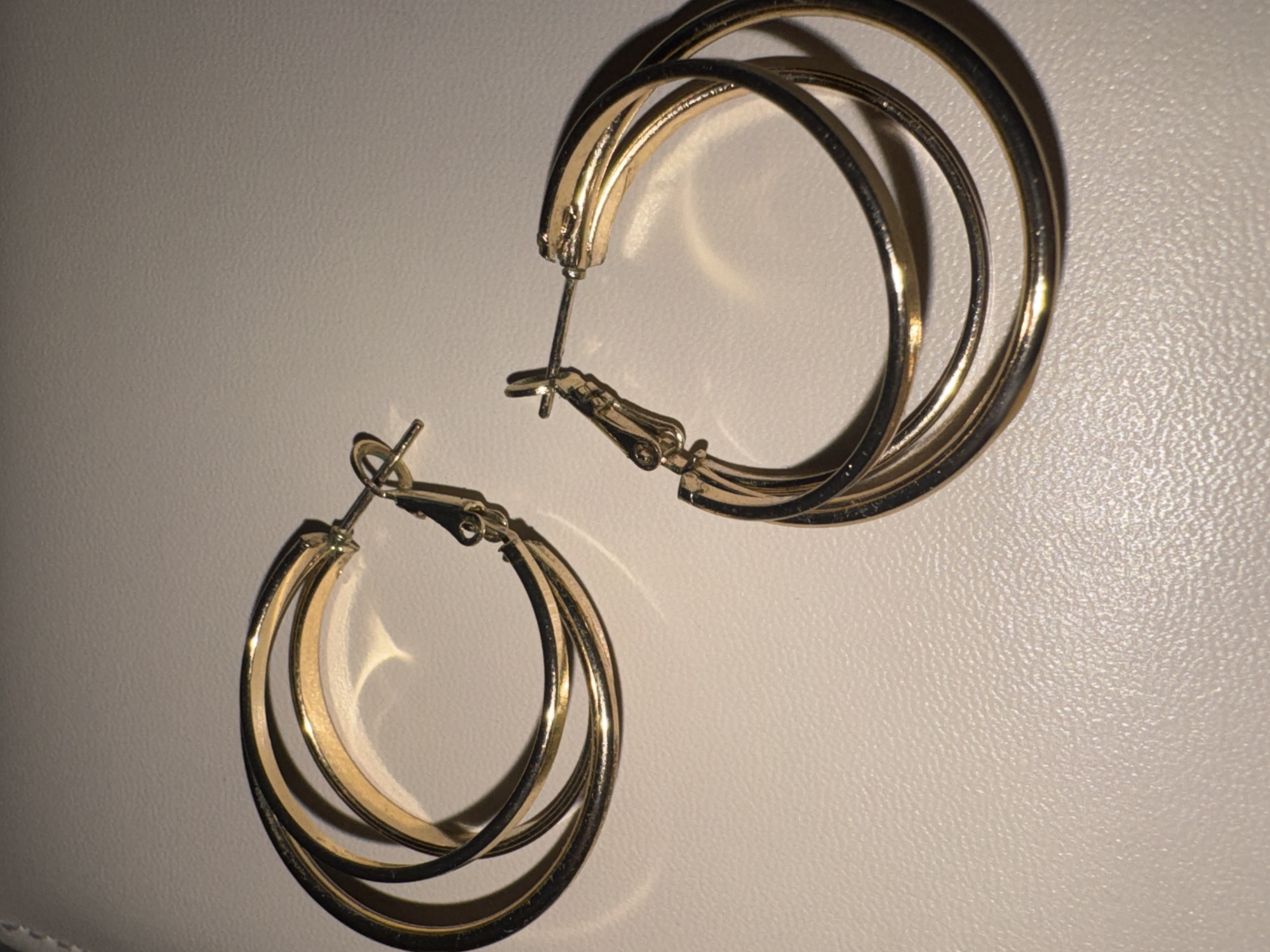 Two gold-colored hoop earrings are laid out on a textured, off-white surface. The earrings are identical, each composed of three concentric hoops. The left earring is positioned towards the top left of the frame, while the right earring is positioned towards the bottom right. The surface appears to be a material like faux leather or a similar textured fabric, commonly found in jewelry displays or on tabletops. There are no people or other objects present in the image. The lighting casts shadows that highlight the curvature and texture of the earrings and the surface. The overall scene is simple and focused on the jewelry. The image does not contain any information that would allow for the determination of its location in the city of Яромінскі сельскі Савет, Belarus.