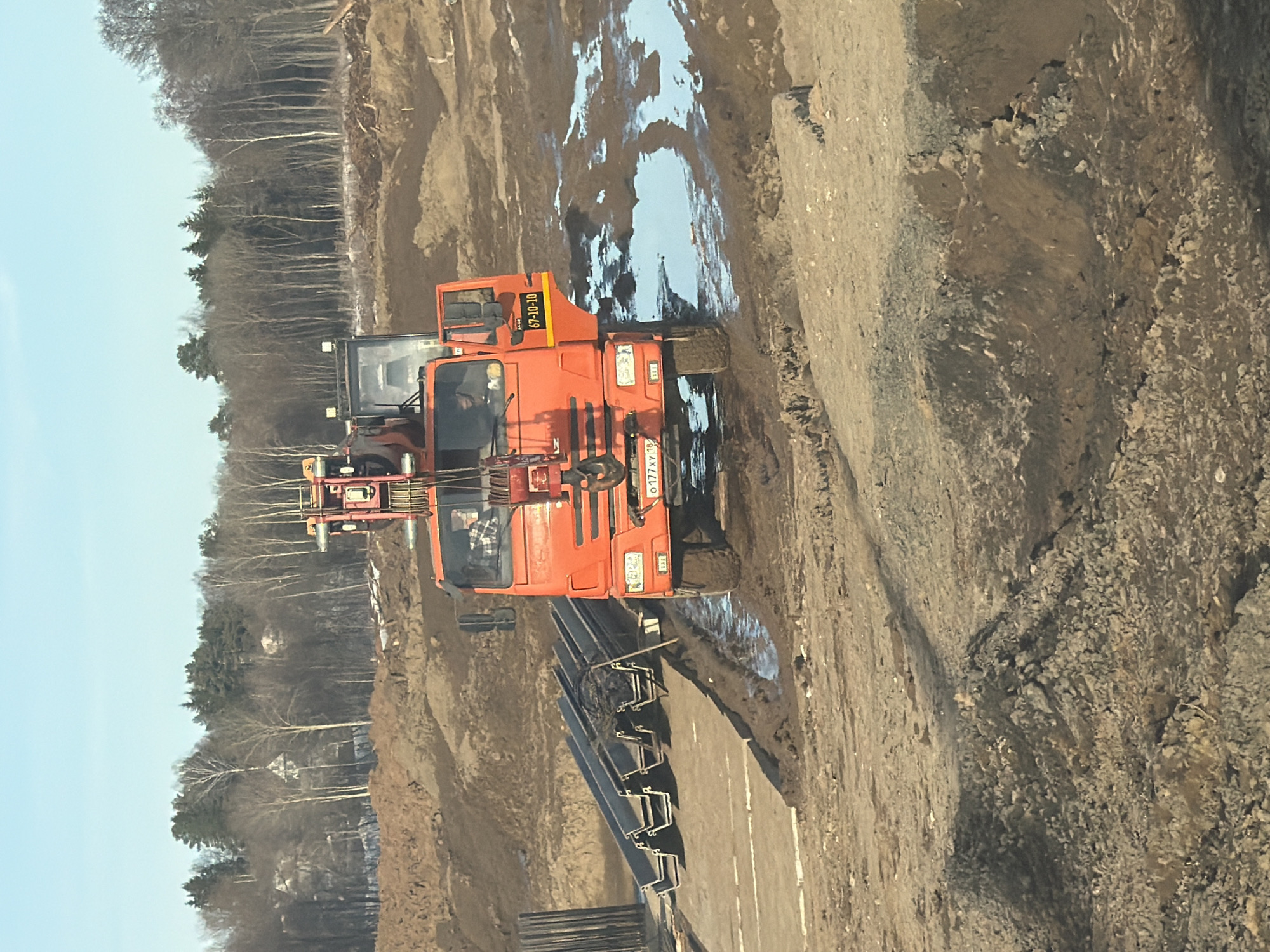 The scene depicts an outdoor construction site in Kabanikha, Russia, under a clear blue sky. An orange heavy-duty truck, equipped with a rear-mounted crane mechanism, is positioned centrally, facing towards the viewer. The truck is heavily soiled with mud on its tires and lower body. Its front license plate, displaying "O 177 XY 18", is clearly visible. The driver's side door is closed, while the passenger's side door is open, revealing a yellow sticker with the sequence "67-10-10". One individual, wearing a plaid shirt, is visible seated within the driver's cabin. To the left of the truck, several dark, elongated metal profiles, resembling C-channels or sheet piles, are stacked horizontally on the ground. The surrounding terrain is a mix of excavated earth, dirt mounds, and significant puddles of water reflecting the sky. In the background, a dense line of deciduous trees, largely bare, interspersed with evergreen conifers, forms a forest edge under the distant horizon.