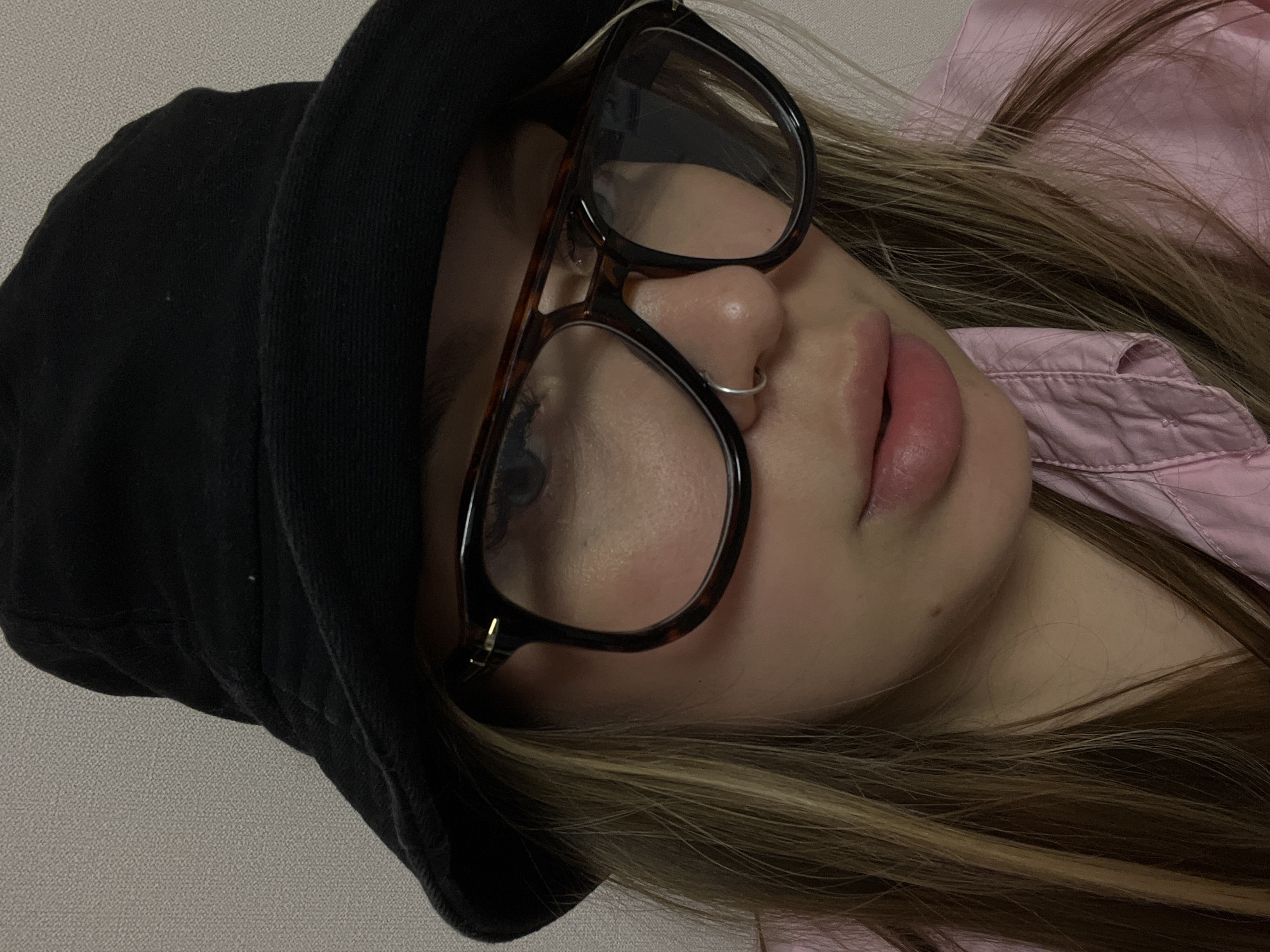 This close-up portrait, taken in Krasnodar, Russia, features a person, likely female, positioned at a tilted angle, possibly lying down or reclining. The subject's face is viewed from the nose down, with the left eye partially visible behind glasses. They are wearing a black bucket hat, which covers the upper part of their head. Large, tortoiseshell-patterned eyeglasses rest on their nose, partially obscuring the eyes. A small, thin silver ring adorns the right nostril.
The person's lips are full and slightly parted, suggesting a relaxed or contemplative expression. Long, streaky blonde and light brown hair cascades over their shoulders, blending with a light pink collared shirt. The background consists of a plain, light-colored textured wall, indicating an indoor setting. The lighting is soft and ambient, without strong shadows, implying an indoor environment rather than a specific time of day. There is no visible text or explicit activity taking place, and the image conveys a calm and candid moment.