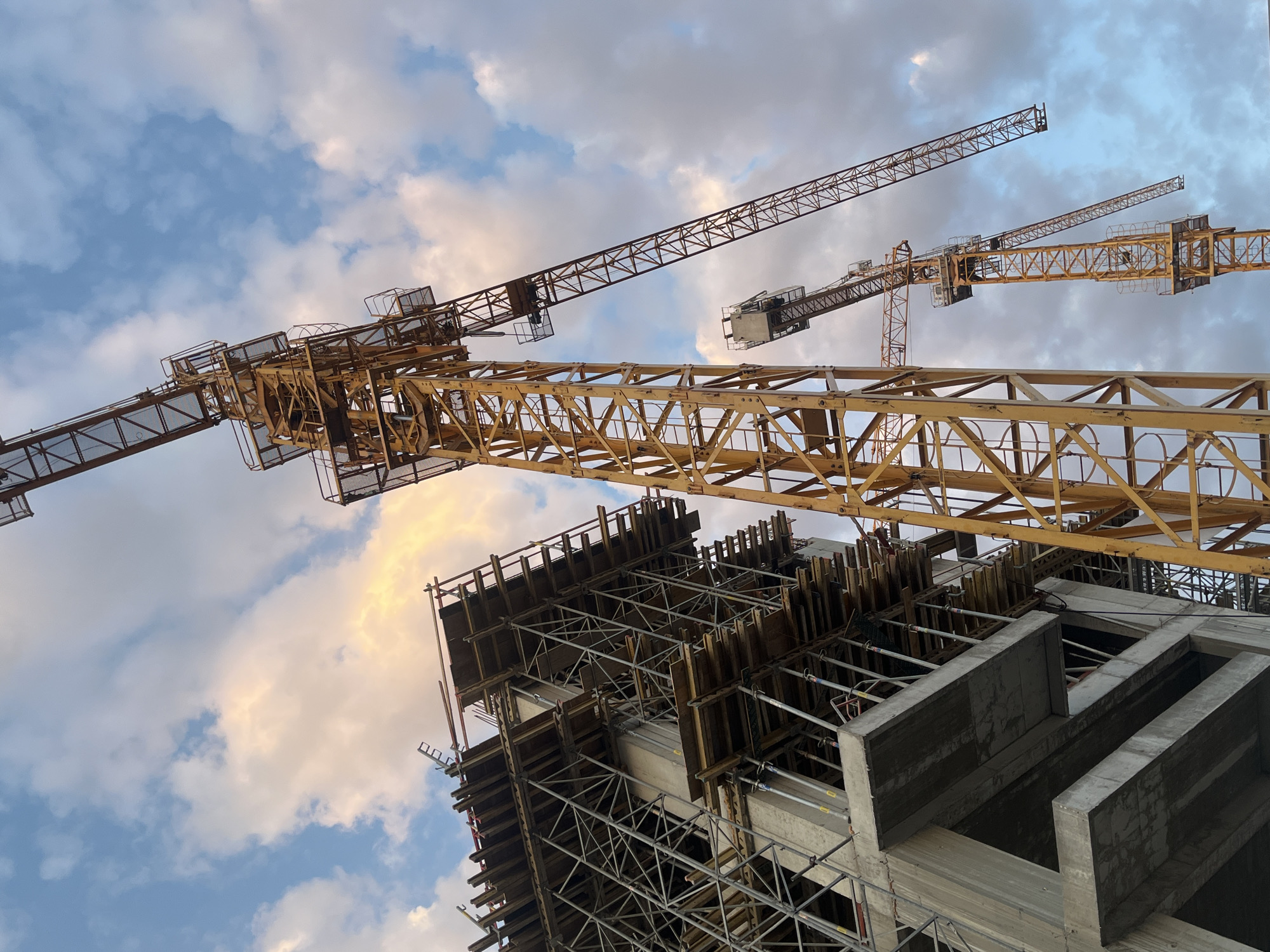 The image depicts a construction site in Belgrade, Serbia, featuring multiple yellow tower cranes against a sky filled with dramatic clouds, illuminated by the setting sun. In the foreground and to the left, a partially constructed building with concrete floors and scaffolding is visible. To the right, a tall tower crane dominates the frame, its boom extending diagonally upwards. Further back and to the right, another tower crane is partially visible, also in yellow. The sky exhibits a blend of blue and soft orange hues, suggesting late afternoon or early evening light. There are no visible people or vehicles in the image, focusing solely on the architectural and mechanical elements of the construction project.
