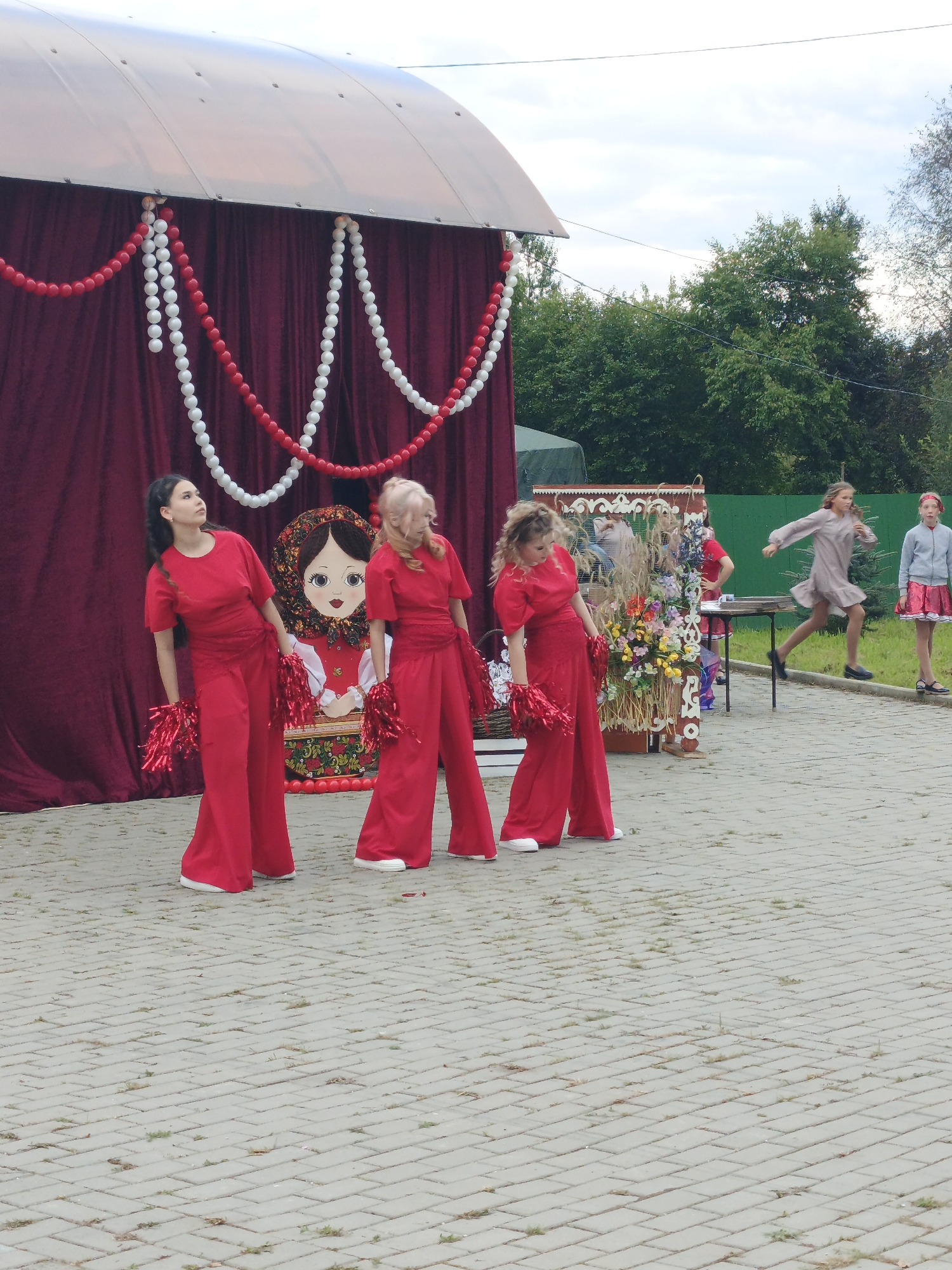 The image depicts an outdoor event in Medvedyovka, Russia, where a group of young women in red outfits are performing. Three women are prominently featured in the foreground, wearing matching red jumpsuits and holding red pom-poms. They appear to be engaged in a synchronized dance or cheerleading routine, with their poses suggesting movement and energy. Behind them, a large, decorated matryoshka doll stands as a backdrop, hinting at a cultural or folk theme for the event. Further in the background, other individuals, including two girls in different attire, are visible, suggesting a broader gathering. The environment consists of a paved area, a covered stage or booth structure draped with red fabric and decorated with red and white bead garlands, and lush green trees in the background under a cloudy sky. The overall scene suggests a festive community event.