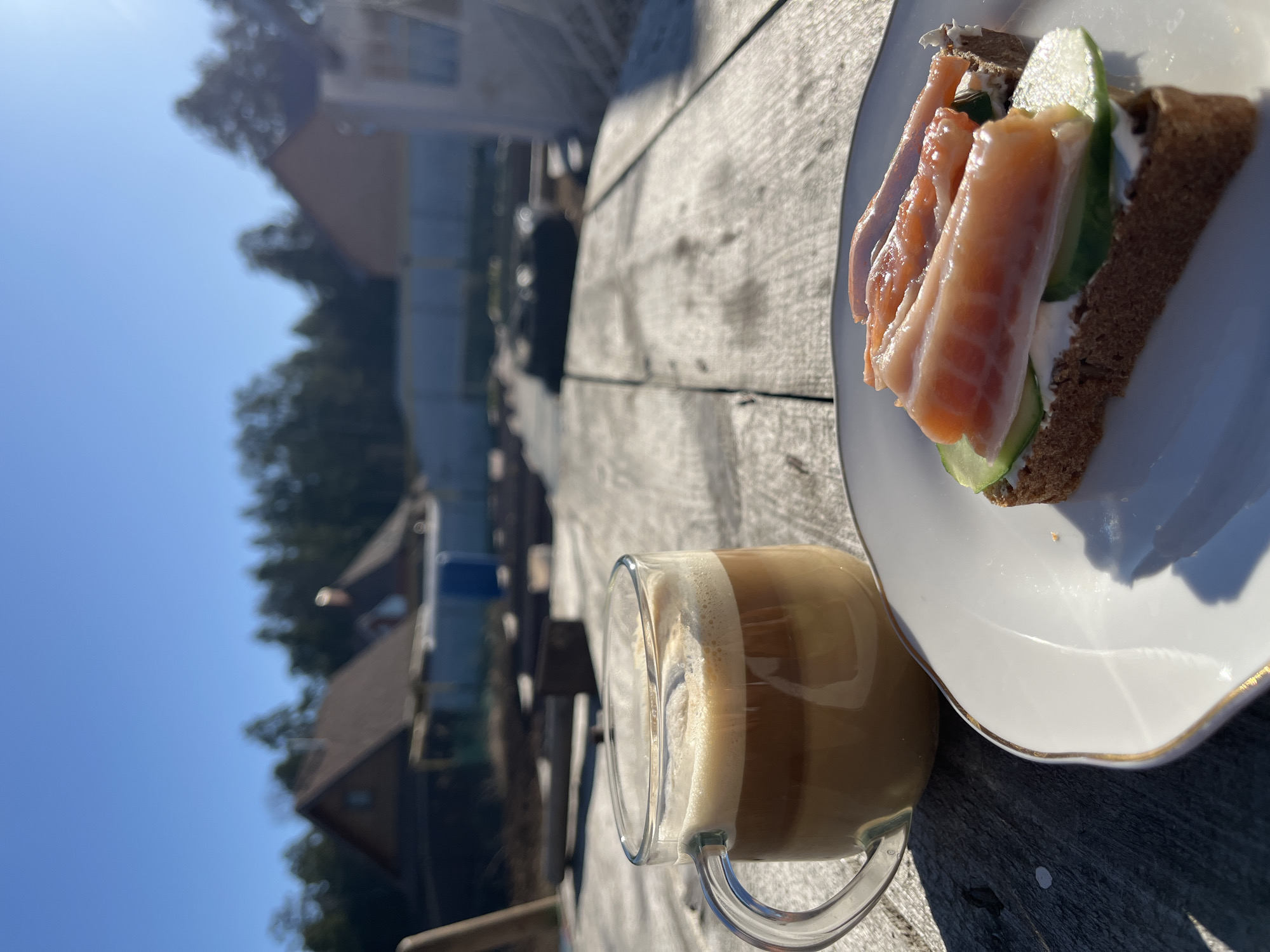 A cup of coffee and an open-faced sandwich rest on a weathered wooden surface outdoors. The sandwich appears to be on dark bread with a creamy spread, cucumber slices, and smoked salmon. It is served on a white plate with a gold trim. The coffee is in a clear glass mug, showcasing layers of coffee and frothy milk. The background is blurred, showing trees, building roofs, and a clear blue sky. The lighting suggests it's daytime. There is no evidence to support the claim that the location is in Chaikovsky, Russia.