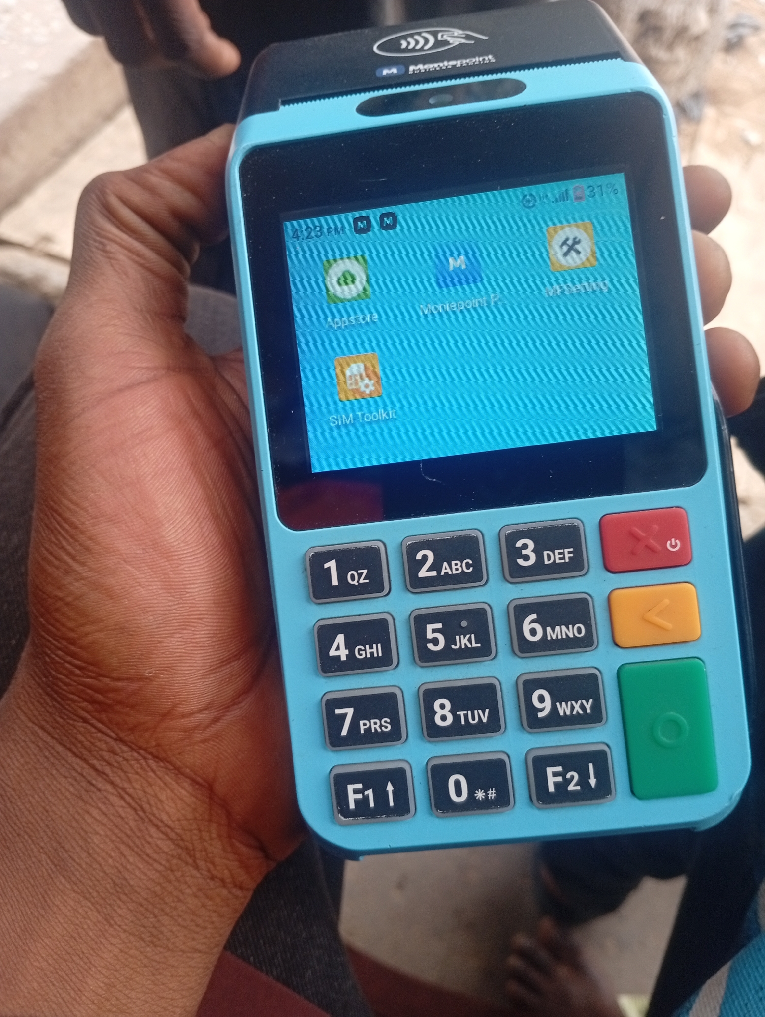 A dark-skinned hand is holding a blue Moniepoint point-of-sale device. The device screen displays icons for "Appstore," "Moniepoint P," "MFSetting," and "SIM Toolkit." The time on the device is 4:23 PM, and the battery is at 31%. The keypad is visible, with buttons for numbers 1-9, 0, F1, and F2, along with symbols and letters. The red power button, orange backspace button, and green enter button are also visible. The background is slightly blurred, showing parts of clothing and feet. The image was taken in Maiburiji, Nigeria.