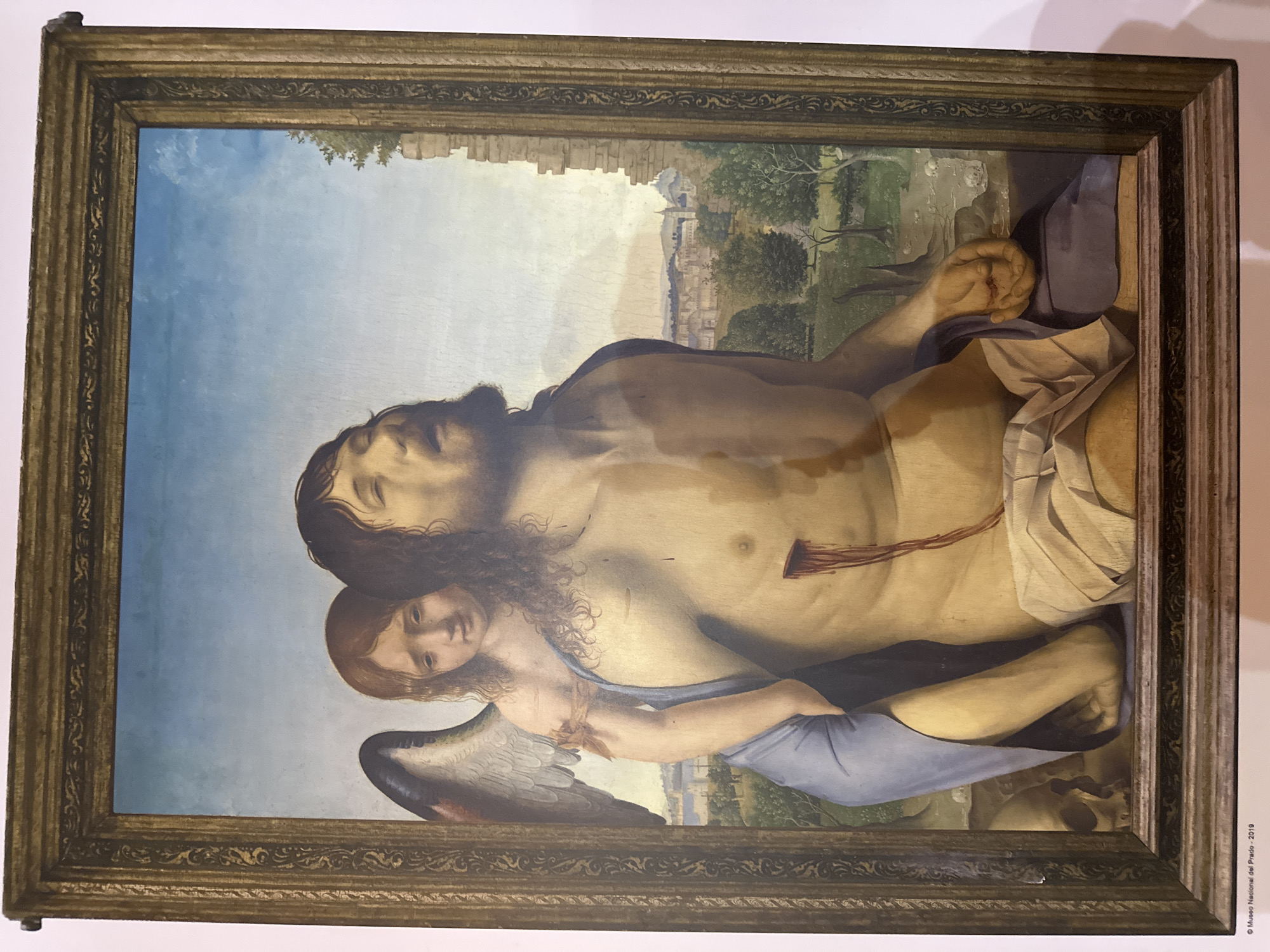 The painting depicts a scene with two central figures. The primary figure is a man, likely representing Christ, with his eyes closed and mouth open, exhibiting signs of suffering. He is depicted shirtless, revealing wounds, notably a prominent wound on his torso with dripping blood. A shroud is draped around his lower body. To his left, a winged figure, an angel, is positioned behind him, partially obscured by the man's body. The angel has a sorrowful expression and is holding the man. The background shows a landscape with trees, rocky formations, and a distant cityscape with architectural structures. The painting is housed in a decorative frame. The lower left corner of the image contains text that reads "© Museo Nacional del Prado - 2019". There is no indication of the city of Mazarrón, Spain, in the image or its associated text.