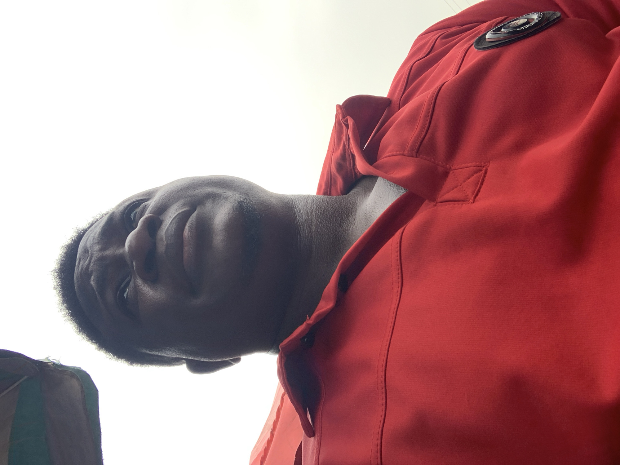 An overhead, low-angle shot captures a person with dark skin, wearing a vibrant red jacket, against an entirely bright white background. The subject's head and upper torso dominate the left side of the frame, looking slightly upward and directly towards the camera with a neutral to contemplative expression.
The red jacket, made of a textured fabric, features visible seams and folds. A circular black and white patch or logo is affixed to the right shoulder/chest area of the jacket, with the word "ORIGINAL" partially discernable on it. The strong upward perspective highlights the person's neck and chin. In the bottom-left corner, a small section of a green and white striped object or fabric is partially visible.
The stark white background suggests either an overexposed sky under bright conditions, indicating daytime, or a brightly lit indoor environment. No specific weather details are evident. The image appears to be a portrait or a selfie, taken from a unique, downward-looking vantage point. While the context given is Akwanga, Nigeria, no specific visual cues within the image itself indicate this location.