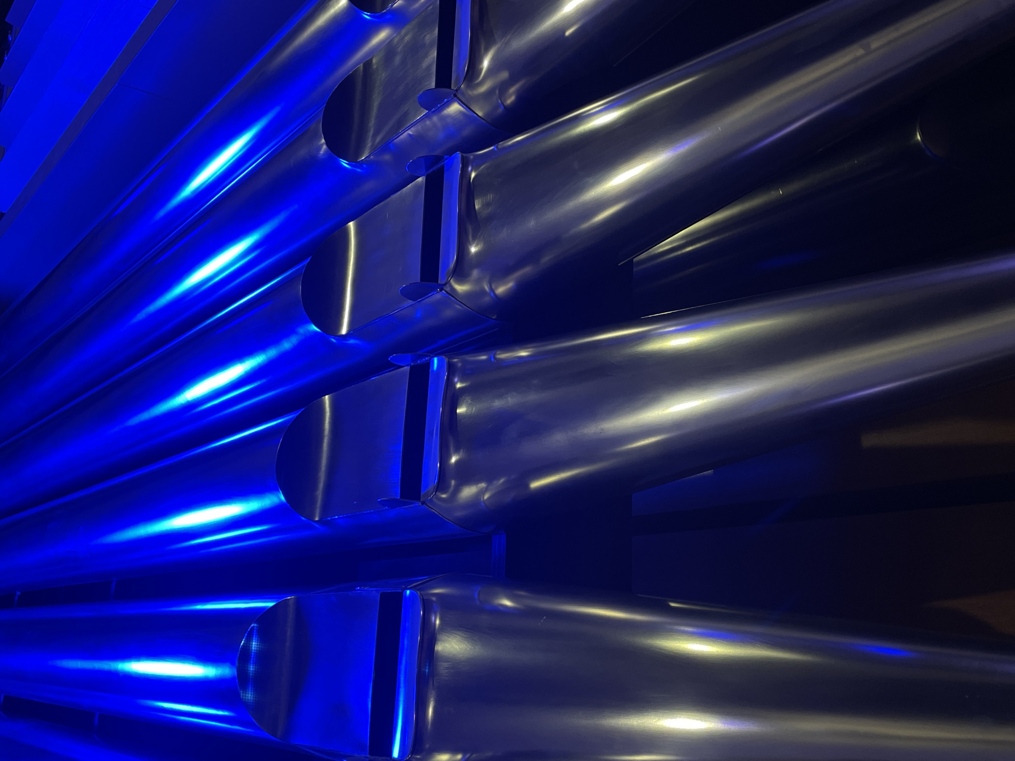 The image presents a close-up, abstract view of numerous large, reflective metallic pipes arranged horizontally in parallel rows. These pipes, characteristic of a grand pipe organ or a contemporary architectural installation, are illuminated by a strong, vibrant blue light originating from the left side of the frame. This intense blue light creates dramatic, elongated reflections and bright highlights along the cylindrical surfaces, making the pipes on the left appear intensely azure.
As the pipes extend towards the right, away from the primary light source, their metallic sheen becomes more apparent, showing a silvery-grey tone with subtle, cooler blue reflections. The dark, almost black background suggests an indoor setting with controlled, theatrical lighting, possibly within a concert hall, church, or public building in Yekaterinburg, Russia. The precise arrangement and varied diameters of the pipes create a sense of depth and intricate design.
No activity or event is taking place; the image is a static study focusing on the structure itself. The striking blue illumination contributes to a cool, modern, and almost ethereal atmosphere. There are no people visible, nor is there any discernible text within the frame. The time of day is indeterminate due to the artificial lighting. The overall impression is one of aesthetic precision and dramatic presentation through the interplay of light, color, and metallic form.