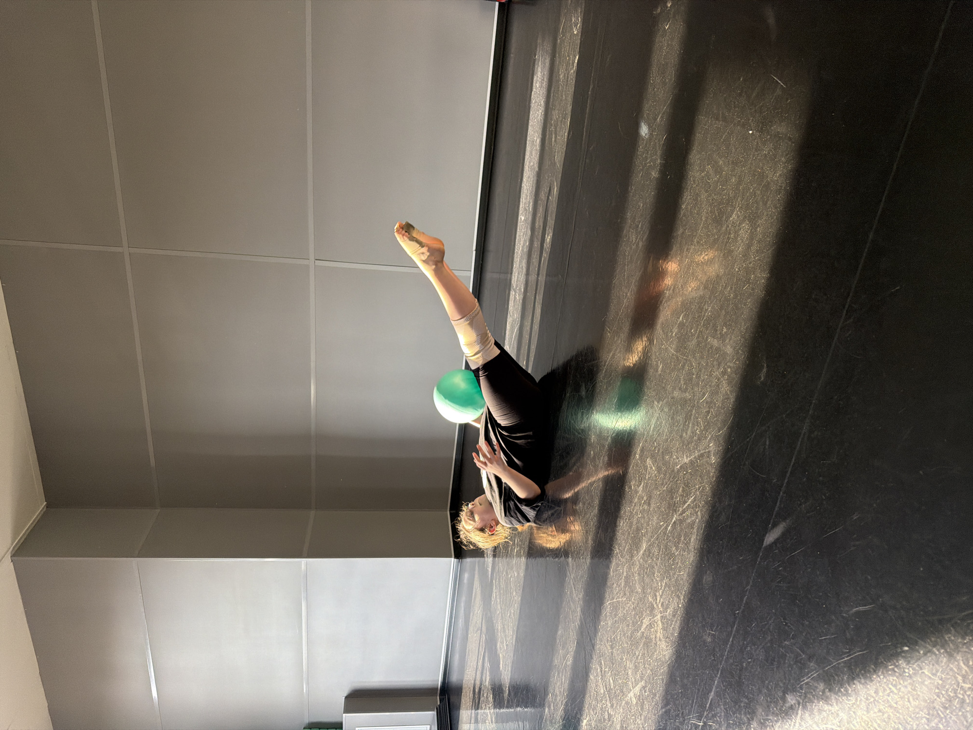 A woman is lying on her back on a dark floor in a room with a gray wall. She has her knees bent and a green balloon is positioned between her legs. Her left leg is raised up. She is wearing black clothing and has light hair. The floor appears to be scratched and there is light shining on it. There is no indication of this being Saint Petersburg, Russia.