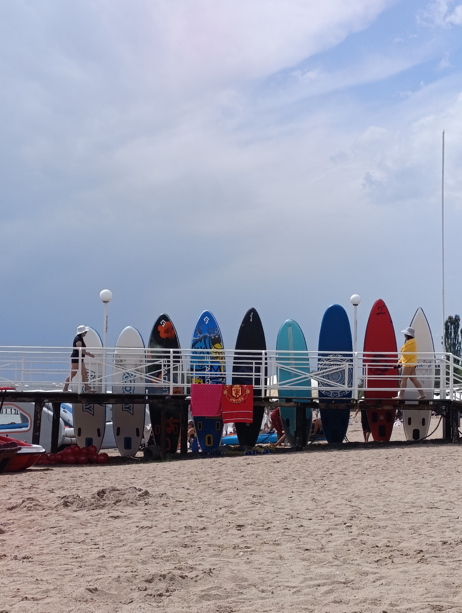 On a sandy beach in Sary-Oy, Kyrgyzstan, a pier is lined with paddleboards and surfboards. A white railing runs along the pier, with a lamp post featuring a white globe light situated between two boards. Several people are visible on the pier and on the sand below. On the pier, two individuals are walking, one wearing a white hat and dark clothing, the other a yellow jacket and hat. A woman is also partially visible near the end of the pier. Several paddleboards are arranged in a row, some with colorful designs and numbering. Beneath the pier, more paddleboards are stacked, and a red inflatable boat is visible on the left side of the image. The beach is covered in light brown sand. The sky above is overcast with patches of blue.