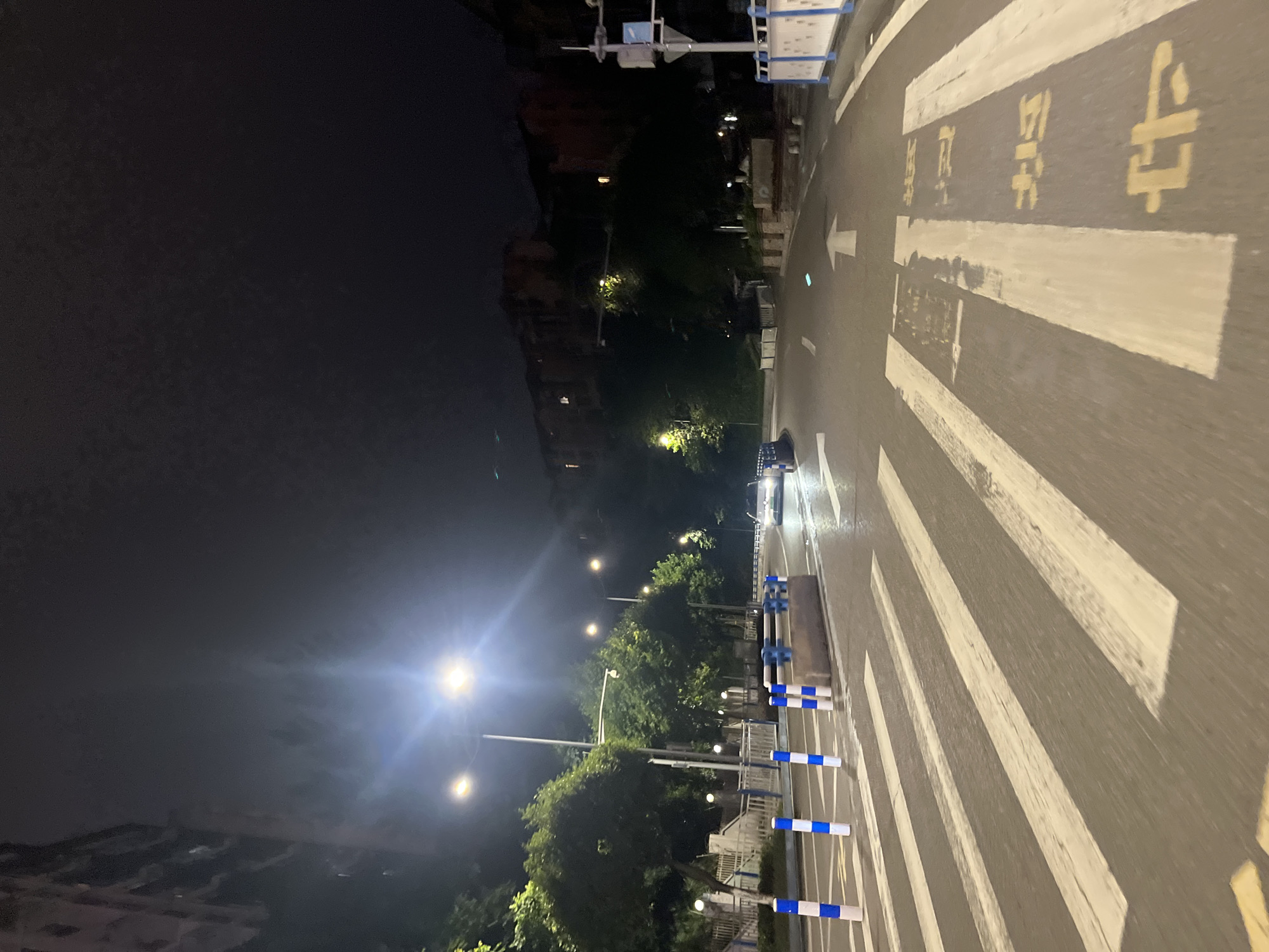 The image depicts a night scene on a wet road in Jiulongpo District, China. The perspective is from a vehicle's point of view, looking forward. A pedestrian crossing with wide white stripes is prominent in the foreground, with yellow painted markings for traffic direction and possibly lane indicators on the asphalt. Blue and white striped bollards are placed along the left side of the road, delineating the pedestrian crossing area. A concrete barrier is visible behind the bollards. Streetlights illuminate the scene, casting bright light and creating reflections on the wet road surface. Trees line the left side of the road, with dense foliage partially obscuring buildings and structures on a hillside in the background. A building with multiple windows is visible on the far left, appearing to be multi-storied. Further down the road, a white car is visible, its headlights on, driving towards the viewer. The road curves gently to the right. On the right side of the road, a traffic light pole with signs and a blue and white barricade with similar bollards are present. The sky is dark, indicating nighttime.