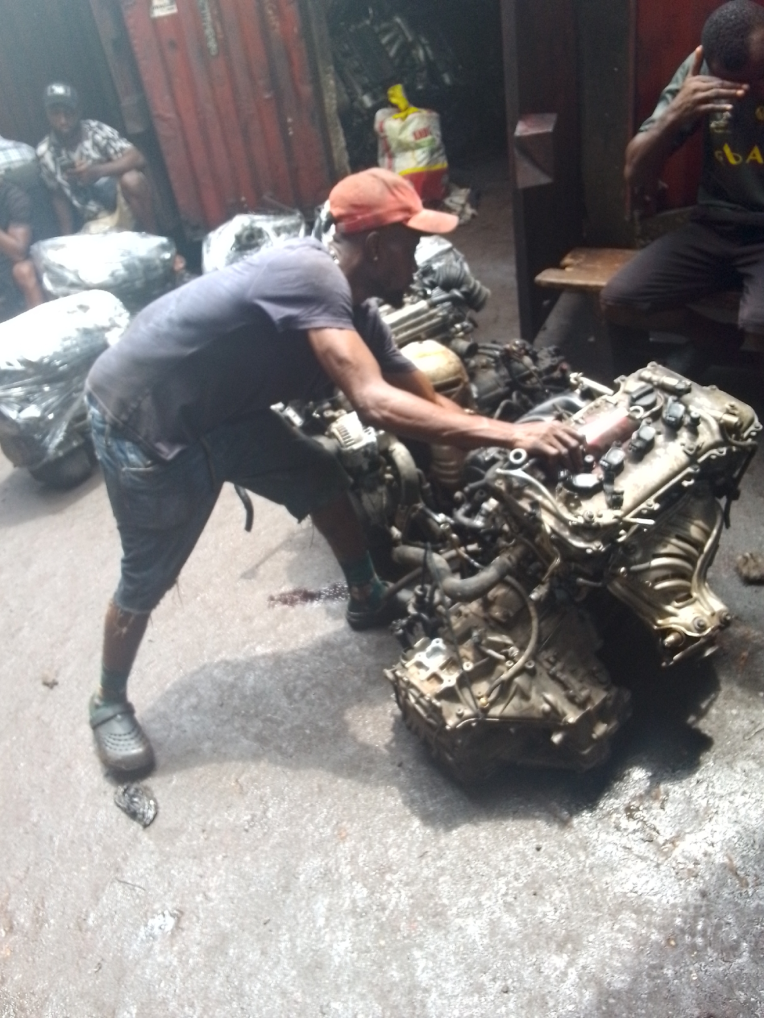 The image shows an outdoor scene in Onitsha, Nigeria, a city known for its bustling markets and automotive parts industry. A man wearing a dark shirt, blue shorts, a red cap, and gray Crocs stands hunched over a large, greasy car engine and transmission, appearing to work on or inspect it. The engine is the prominent subject. Other men are present in the background: one sitting on a bench and another squatting. Wrapped bundles, likely containing automotive parts or components, are seen on the left side of the frame. The ground is concrete and appears oily. The overall setting is industrial.