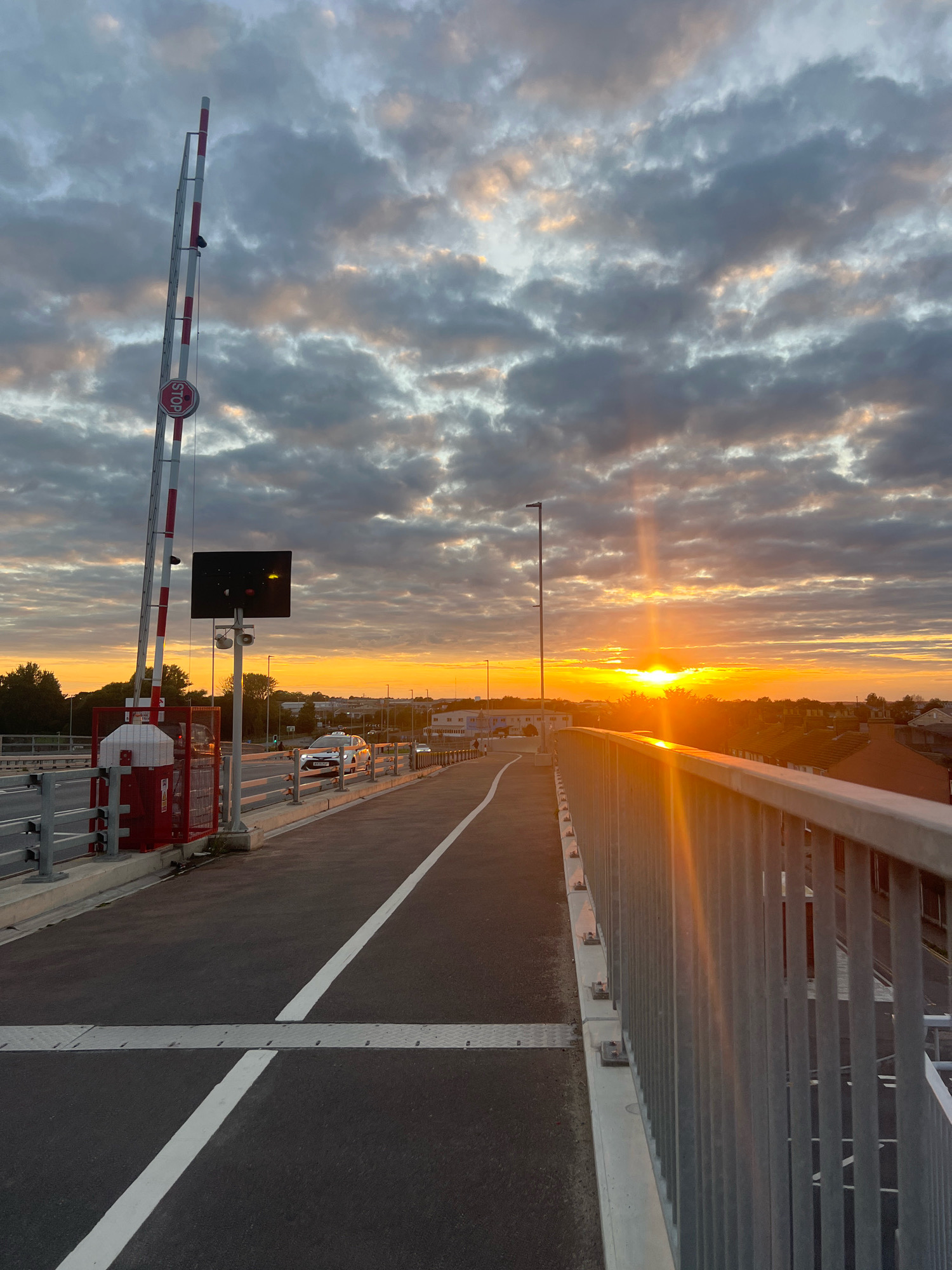 This is a photograph of a bridge in Great Yarmouth, United Kingdom, at sunset. The bridge appears to be a road bridge, with a pedestrian walkway on the right side. A red and white barrier arm is visible on the left side of the bridge, indicating it may be a level crossing or a bridge that can be raised. A traffic light is also visible on the left, showing a yellow light. The sky is filled with dramatic clouds, tinged with orange and pink from the setting sun. The sun itself is low on the horizon, casting long rays of light across the scene. A few cars are visible on the road, and houses with red-tiled roofs can be seen in the background. The overall impression is one of a calm and beautiful end to the day.