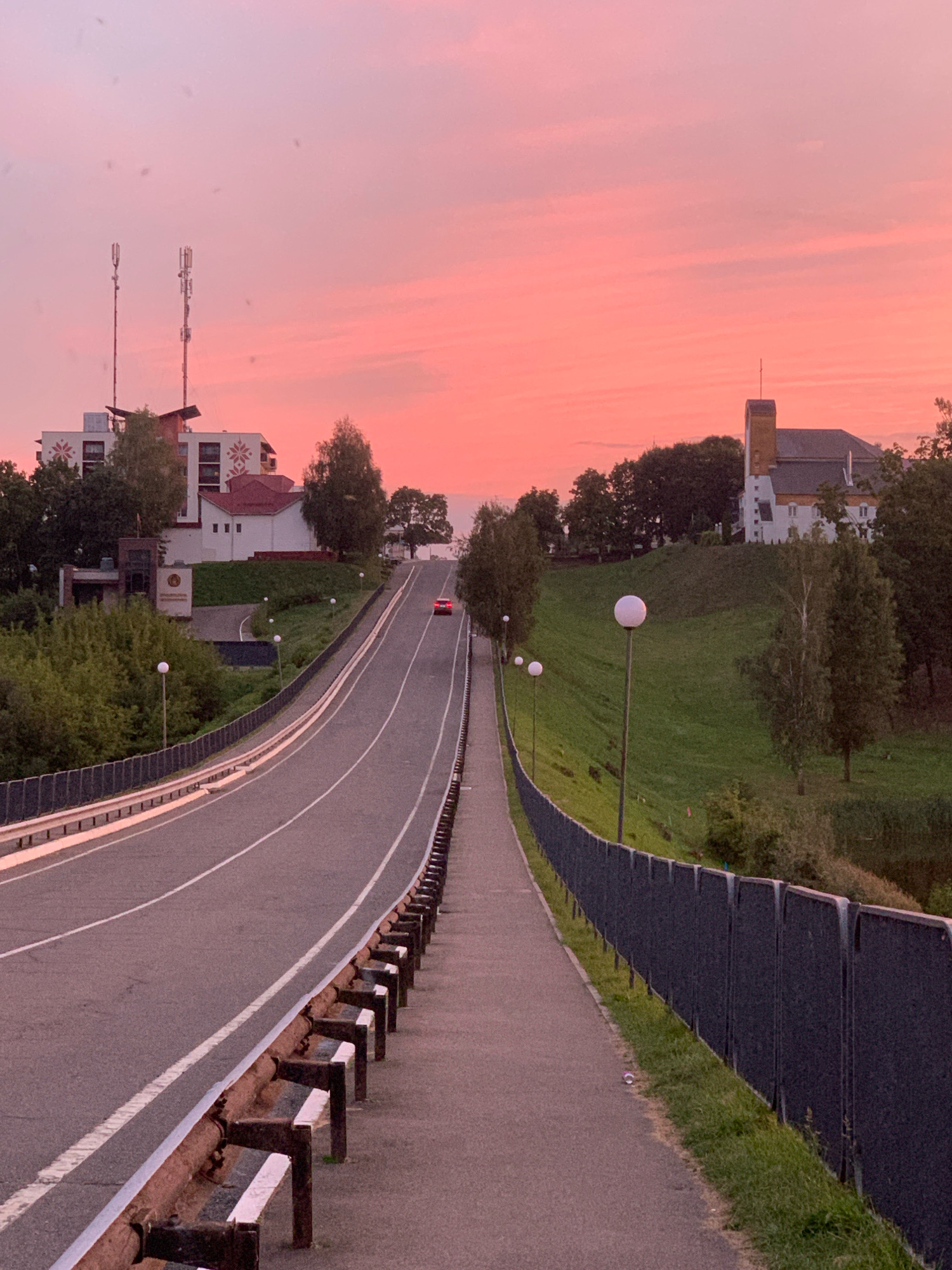 The image shows a street in the city of Kapyĺ, Belarus, during sunset. A red car is driving on the road. The sky is pink, and there are buildings and trees on both sides of the road. There is a pedestrian walkway next to the road, separated by a fence. The road is paved and has white lines marking the lanes. There are streetlights on the walkway.