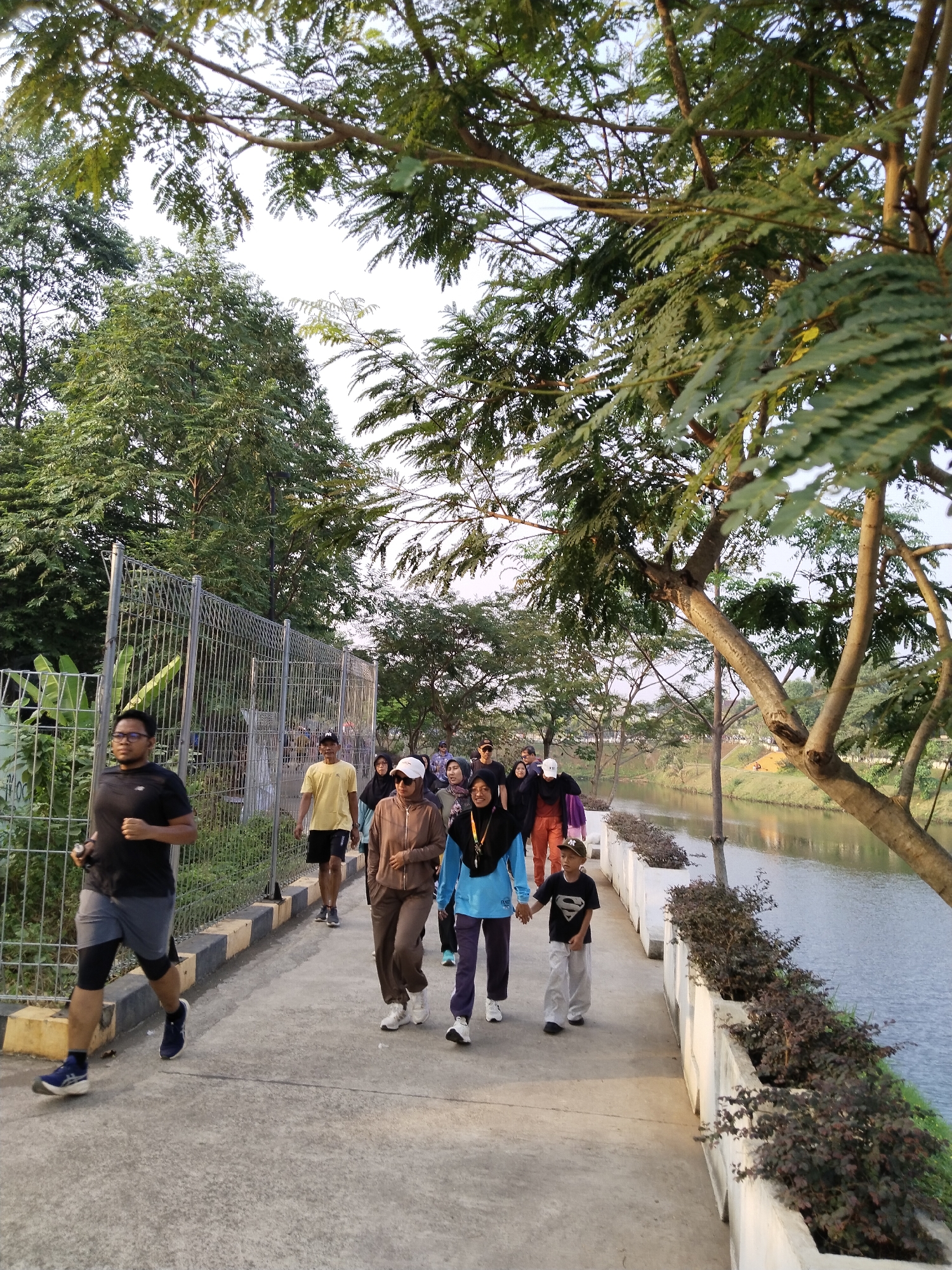 In South Jakarta, Indonesia, a group of people are walking and running along a paved path beside a body of water. The path is lined with a white barrier and planters filled with dark green foliage. A metal fence runs along the left side of the path, separating it from a grassy area with trees. A runner in a black t-shirt and grey shorts leads the group. Behind him, a man in a yellow t-shirt and black shorts walks. Further back, several individuals, including women wearing hijabs, are walking together. A young boy wearing a "Superman" t-shirt walks hand-in-hand with another person. Trees with lush green leaves overhang the path, casting shadows. The sky is visible above the trees, appearing bright and possibly overcast. The overall scene suggests a recreational activity taking place in an urban park setting.