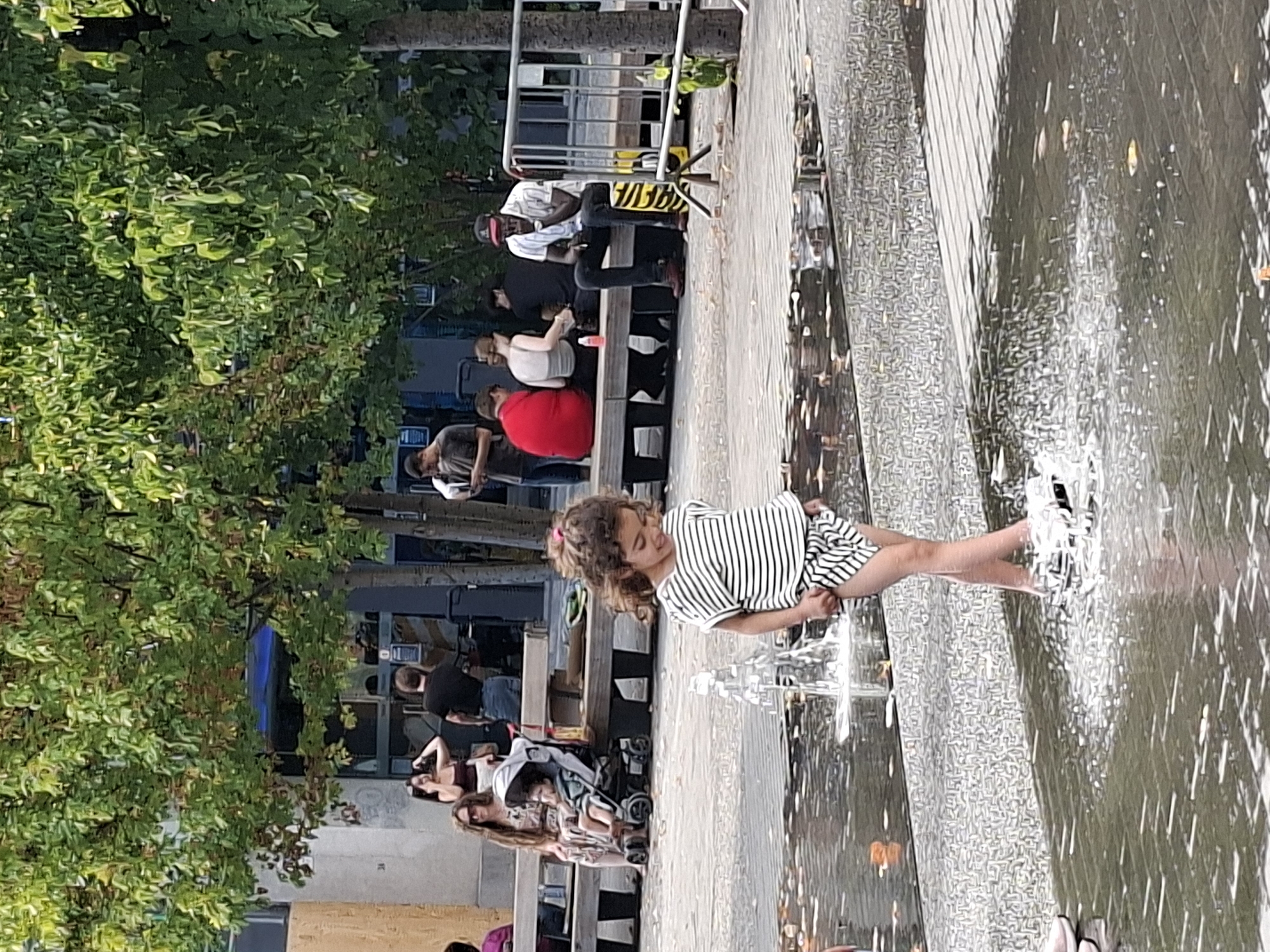 The image captures a lively scene in a public urban space, likely a plaza or park area, with elements suggesting it is located in the city of Brussels, Belgium. In the foreground, a young girl with curly brown hair, wearing a striped t-shirt and shorts, is playfully splashing in a shallow water feature. The water jets create a dynamic spray around her feet, and she appears to be smiling and enjoying herself.
In the mid-ground, a group of people are seated on a long wooden bench. To the left, a woman with long, wavy brown hair and sunglasses is seated with a child in a stroller beside her. Another woman to her right is wearing a patterned dress. Further along the bench, several other individuals are visible, some interacting with each other, while one man in the foreground of this section, wearing a white shirt and a baseball cap, is smoking. The background reveals more of the urban environment, with trees providing shade and a building with windows. A metal fence is also visible on the right side of the image. The overall atmosphere is one of casual public enjoyment, with people relaxing and children playing in a refreshing water display on a sunny day.