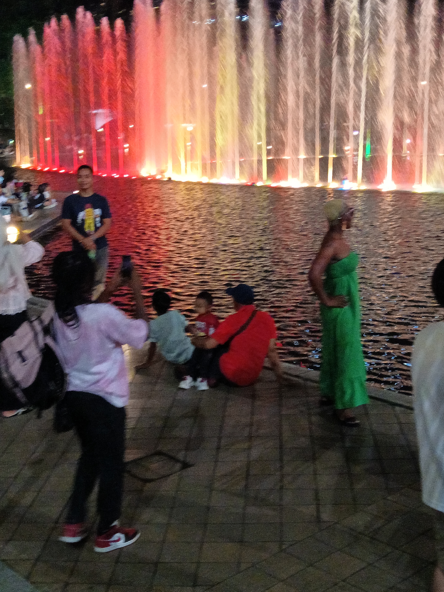 The image depicts a nighttime scene at the Kuala Lumpur City Centre (KLCC) Park, famous for its Symphony Lake water fountain shows. In the foreground, a young woman wearing a pink shirt and black pants is captured from the knees down, as she walks on a paved surface. She is wearing red and white sneakers. To her left, a black backpack is visible. In the mid-ground, a person in a light blue shirt and dark pants is seated, facing away from the camera. Next to them, a child in a red shirt and dark pants is also seated, looking towards the fountains. A woman in a bright green strapless dress stands to the right, facing away from the camera with her hand on her hip. She has a light-colored headscarf. Behind them, a man in a dark t-shirt stands by the water's edge, looking towards the fountains. The background is dominated by the illuminated water jets of the Symphony Lake, with streams of water lit in red, yellow, and white, creating a dynamic display. The water is rippling and reflecting the colorful lights. The overall atmosphere suggests a public gathering, likely a family outing, to enjoy the fountain show at dusk or night.