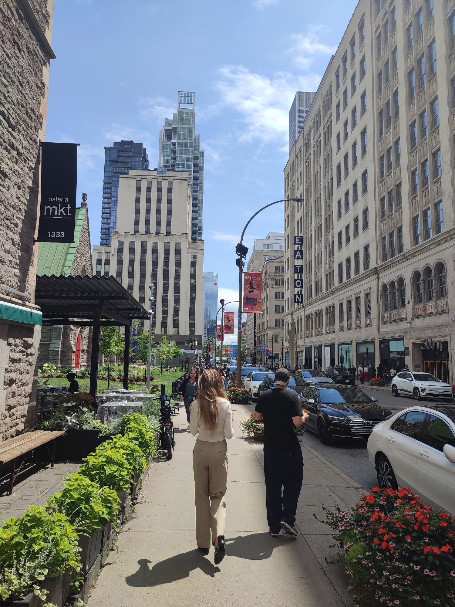 The image depicts a street scene in Montreal, Canada, likely in the downtown area, characterized by a mix of historic and modern architecture. On the left, a stone building with a green sloped roof and a black sign reading "osteria mkt 1333" is visible. Adjacent to it is an outdoor seating area with tables and chairs, bordered by planters filled with lush green foliage. A wooden bench is also present on the sidewalk.
The sidewalk is populated by several individuals. In the foreground, a woman with long blonde hair, wearing a cream-colored sweater and beige pants, walks away from the camera. Beside her, a man in a black t-shirt and dark pants also walks away. Further down the sidewalk, other pedestrians are seen walking in both directions.
The street itself is lined with cars, including a white sedan and a black Audi. The background reveals a cityscape with tall buildings, including a modern skyscraper with a distinctive top structure, and older buildings with numerous windows. Red and white banners hang from lampposts on the right side of the street, and a large sign with vertical letters spelling "EATON" is visible on one of the buildings. The sky is blue with scattered clouds, suggesting it is daytime. The scene conveys a vibrant urban atmosphere.