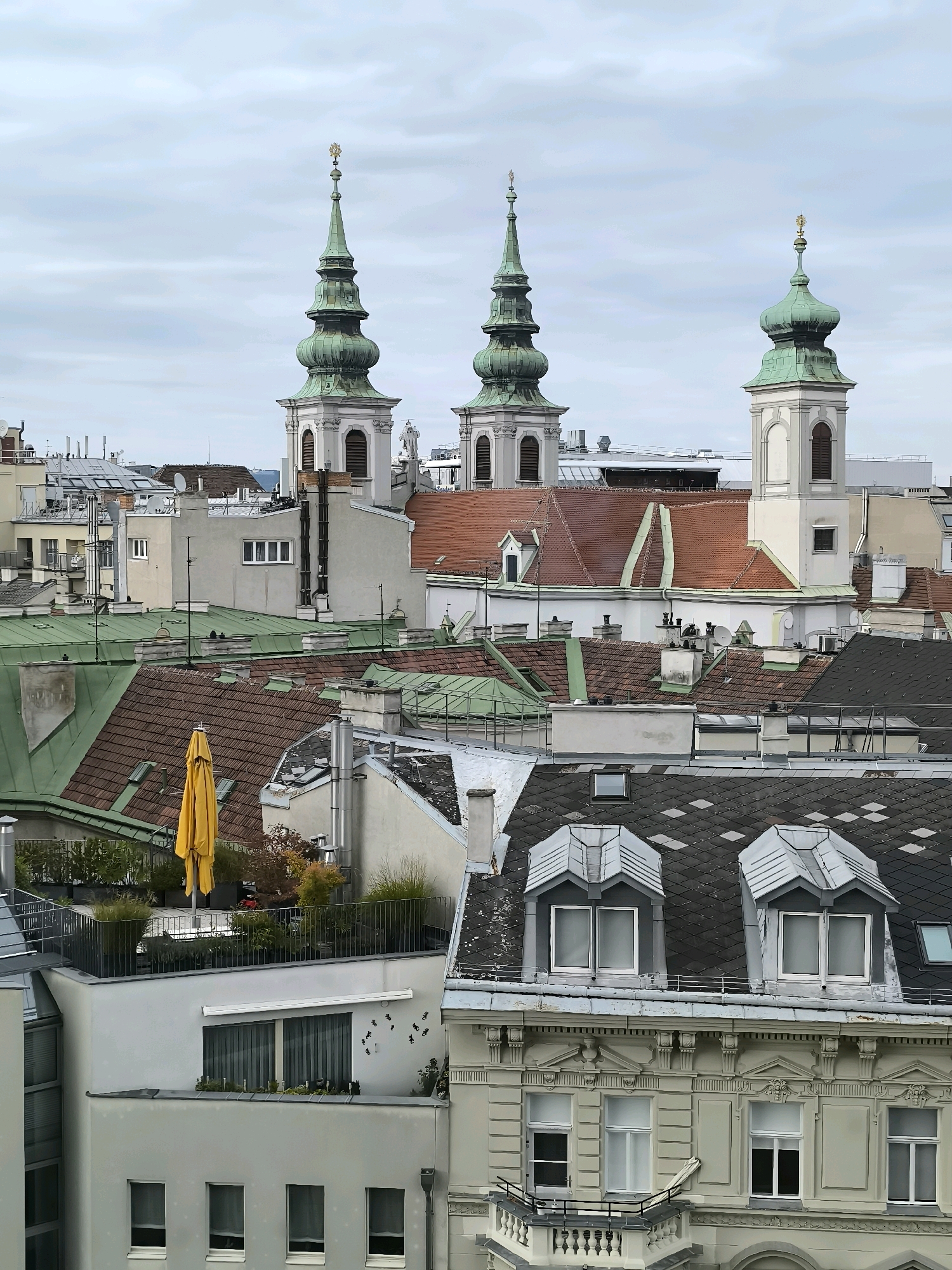 An exterior, high-angle, eye-level medium shot captures a cityscape with a cloudy sky. In the center of the frame, three church spires with green roofs and gold crosses rise above a dense collection of buildings. The church in the middle is flanked by two others, one to its left and one to its right, all exhibiting similar architectural styles. The surrounding buildings feature a variety of roof types, including red-tiled, green metal, and dark grey shingles. Some buildings have visible chimneys and dormer windows. In the foreground, a multi-story building with ornate architectural details and several windows is visible. A balcony with a black railing juts out from this building. A rooftop terrace with greenery, a yellow umbrella, and outdoor seating is also present on one of the buildings. The overall impression is one of a densely populated urban environment with a mix of historical and modern architecture.
