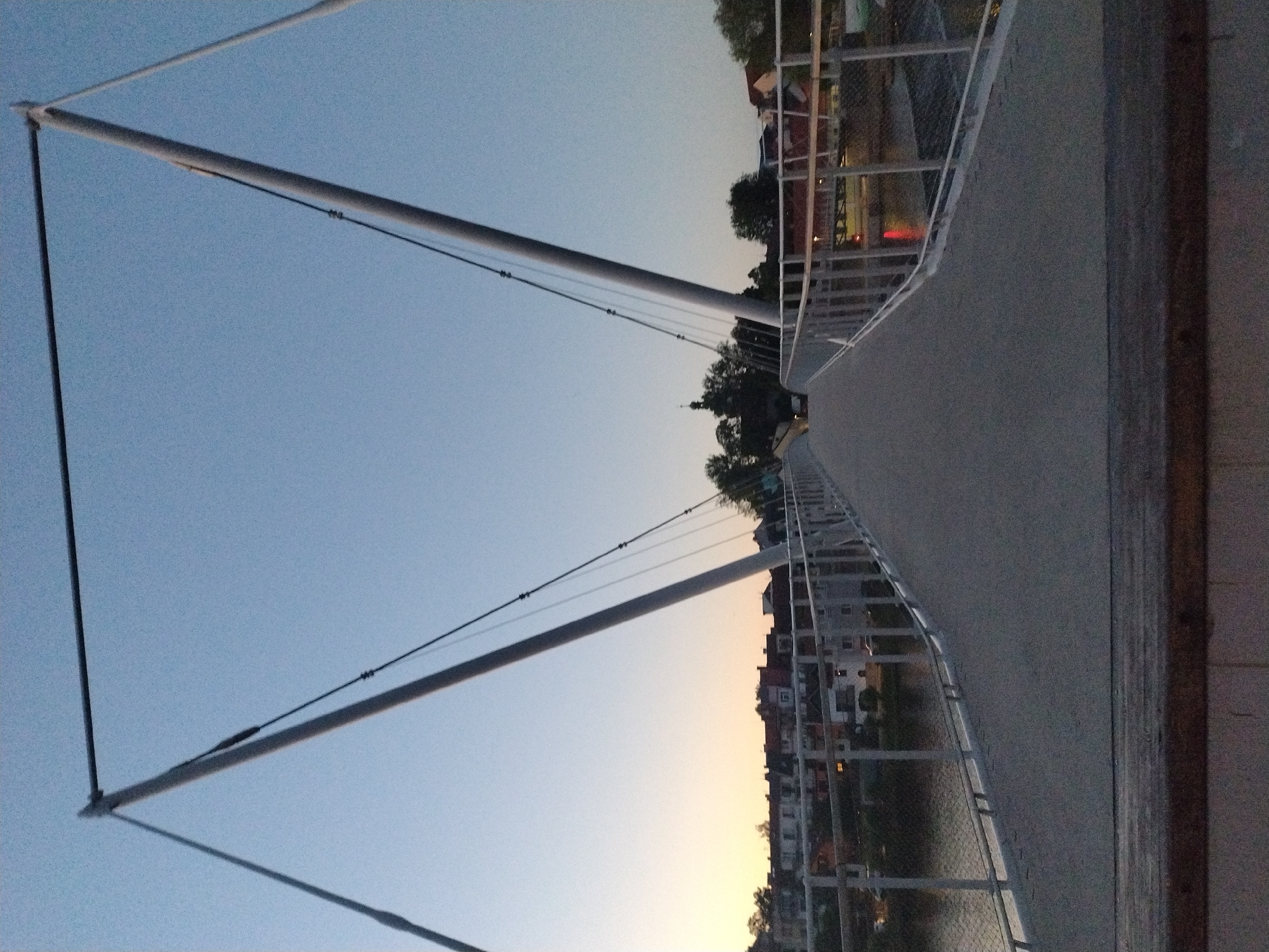 The image captures a pedestrian bridge in Písek, Czechia, at dusk. The bridge, with its modern, minimalist design, features two tall, angled pylons connected by a horizontal bar at the top, from which cables descend to support the walkway. The walkway itself is a gray, concrete surface with white metal railings on either side. In the background, the town of Písek is visible, with buildings lining the riverbank and a church steeple rising above the trees. The sky is a clear, pale blue, transitioning to a warmer hue near the horizon, indicating the setting sun. The river flows gently below the bridge, reflecting the dim light. No people or vehicles are clearly discernible in the scene. The overall impression is one of tranquility and urban landscape at the close of day.