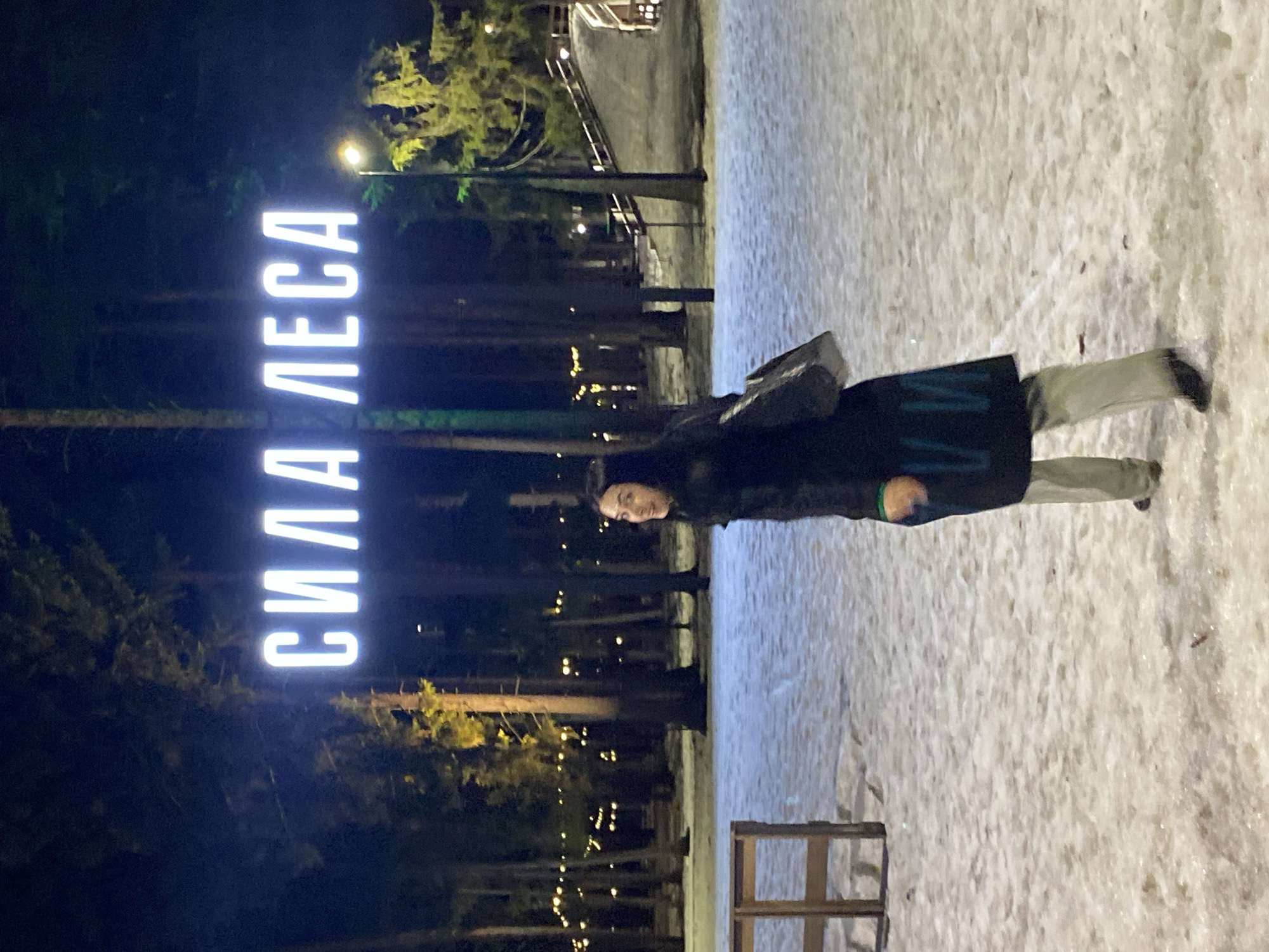 A medium shot depicts a person standing in a snowy area next to a lit sign reading "СИЛА ЛЕСА". The scene appears to be at night, in Votkinsk, Russia, near a forested area. The sign's vertical orientation is illuminated, contrasting against the dark trees in the background. The person is wearing a dark coat and light-colored pants, positioned to the right of the sign. A wooden structure is visible in the lower right foreground, adding to the outdoor setting. The ground is covered in snow, reflecting light and adding texture to the scene. Street lights and other light sources are visible in the background, contributing to the ambient lighting.