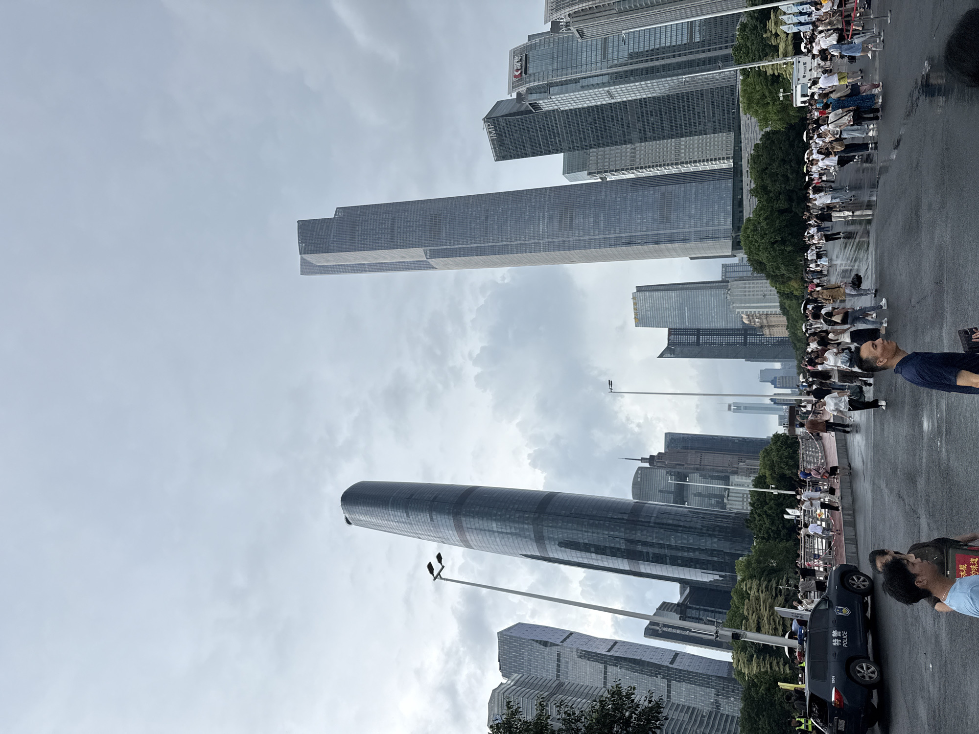 The image captures a wide-angle view of a street scene in the Tianhe District of China, characterized by a dense collection of modern skyscrapers. The sky is overcast with dramatic clouds, casting a diffused light over the urban landscape. In the foreground, a wet, paved road suggests recent rainfall, with a large crowd of people moving about. Several individuals are captured in mid-stride, some holding umbrellas, while others are standing in groups.
Dominating the mid-ground and background are towering glass and steel buildings of various architectural designs. The most prominent are two exceptionally tall, slender skyscrapers, one with a curved, tapering form and the other with a more rectangular, stepped profile. To the left of the curved skyscraper, a police vehicle, marked with "POLICE," is parked. Streetlights are visible along the roadway, and trees line the edges of the scene, separating the street from the buildings. The overall atmosphere appears to be one of public gathering or pedestrian traffic in a bustling commercial district.