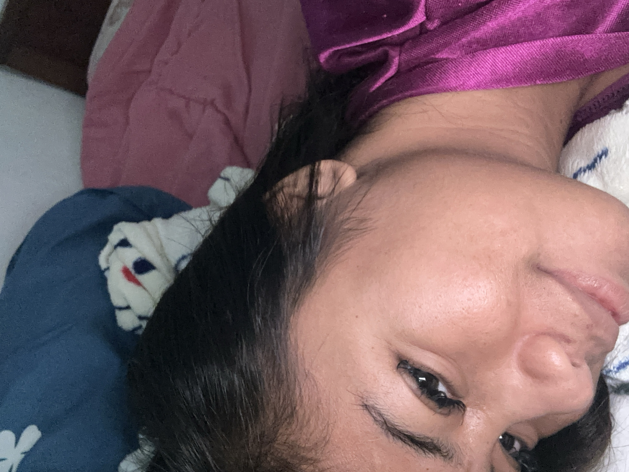 A young woman is lying on her side on a bed, looking up at the camera. She has dark hair and is wearing a purple top. The background is a mix of pink and blue bedding. The image is a close-up and slightly blurry, suggesting it was taken with a mobile phone. The context of Sukawati, Indonesia, implies a warm, possibly humid environment, and the bedding suggests an indoor domestic setting.