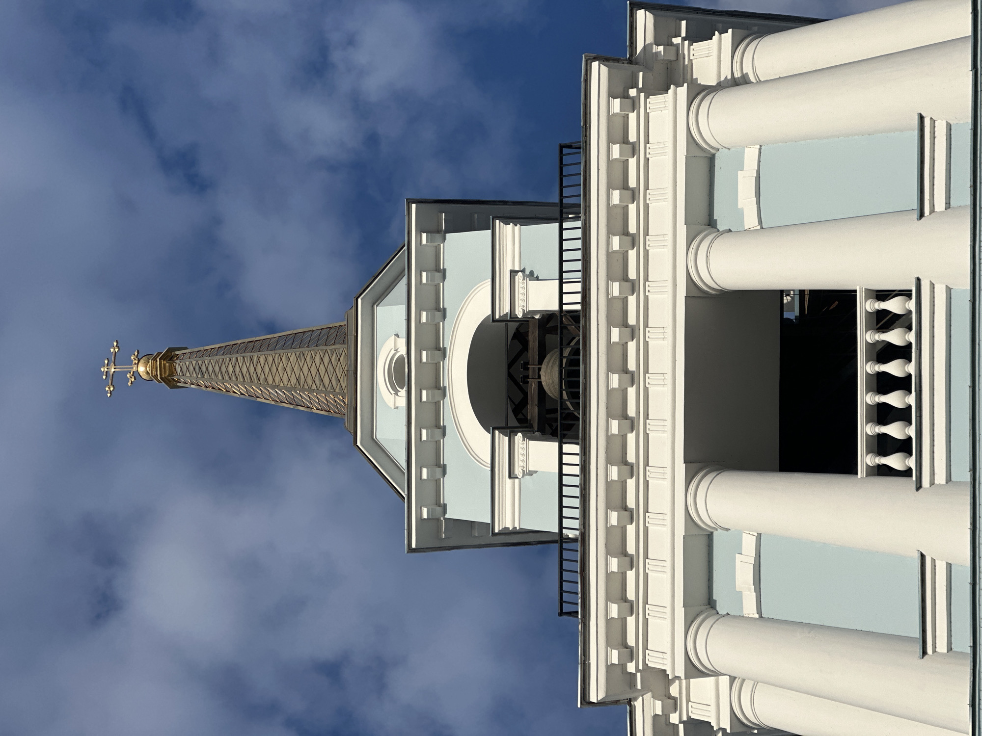 The image shows a portion of the Holy Trinity Cathedral in Votkinsk, Russia, viewed from a low angle. The architectural details of the cathedral are visible, including white columns, light blue walls, and a golden spire topped with a cross. The sky behind the cathedral is blue with scattered clouds.